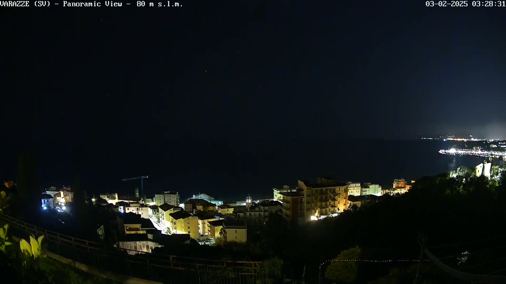 A nighttime panoramic view of a coastal town, illuminated by lights, with a dark sea extending to a dimly lit shoreline in the distance under a clear, starry sky.