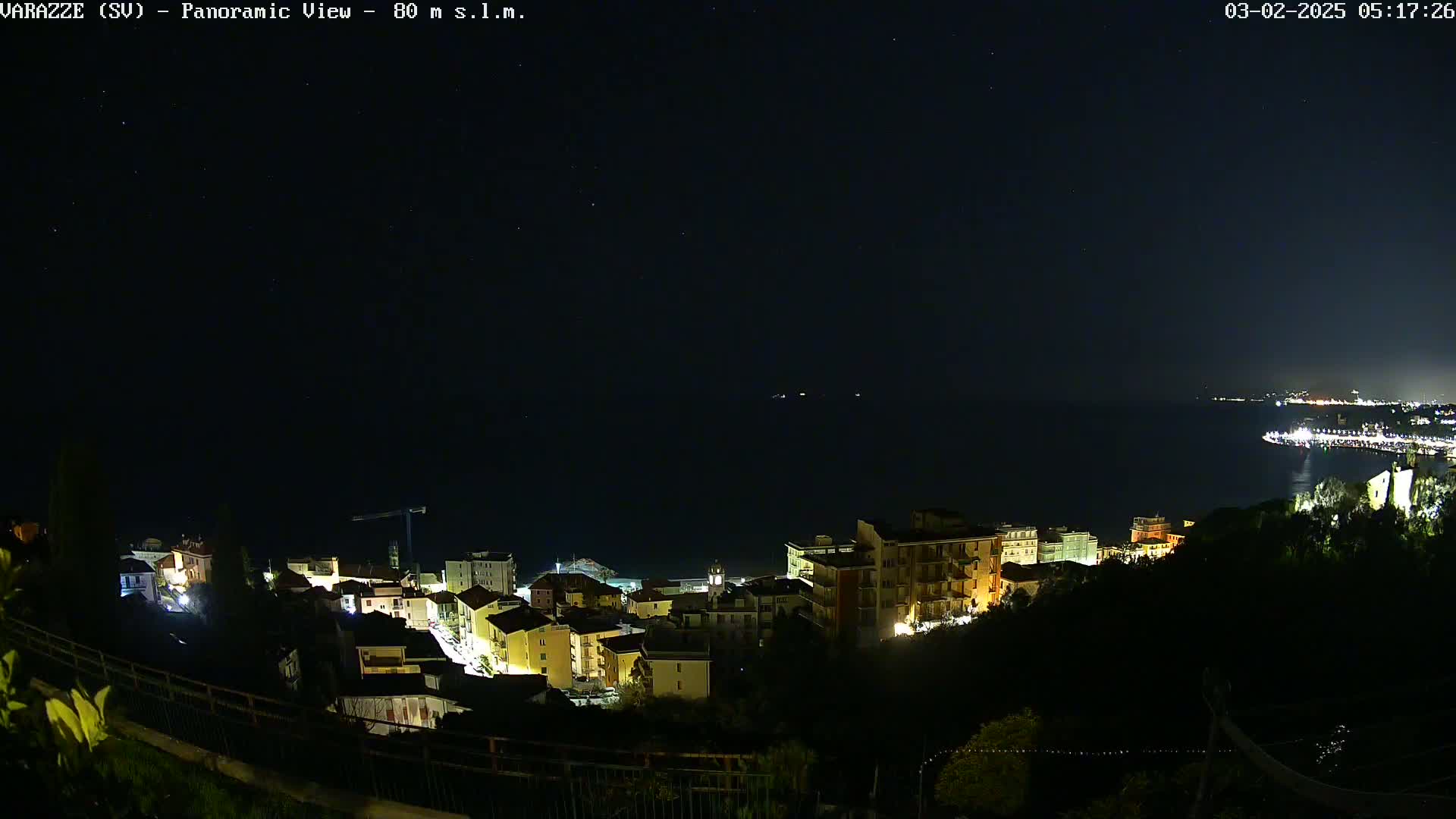 A nighttime panoramic view shows a coastal town illuminated, with a dark sea and starry sky in the background.