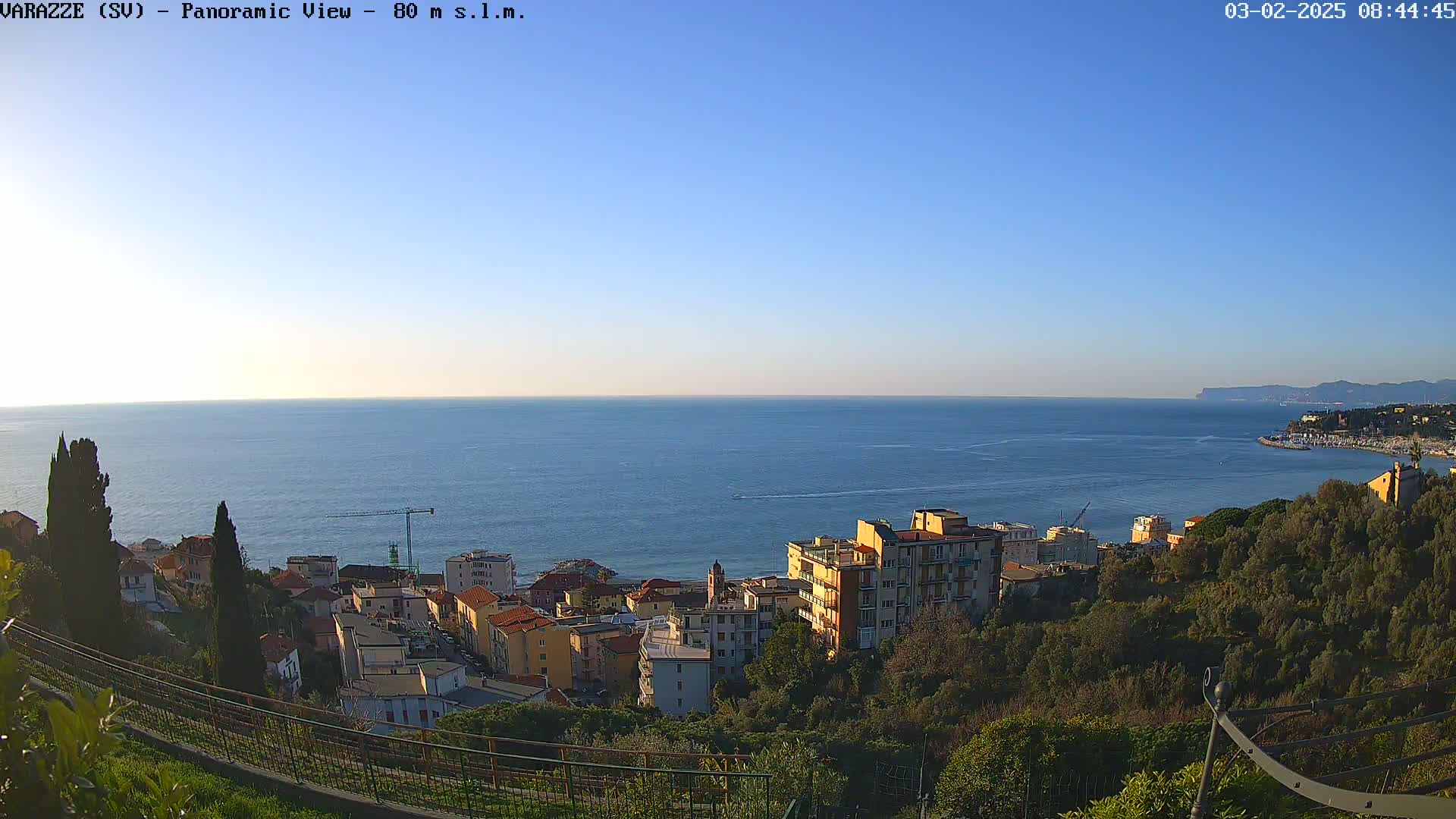 A panoramic view of a coastal town on a clear, sunny day shows a calm ocean, buildings nestled on a hillside, and lush green vegetation.