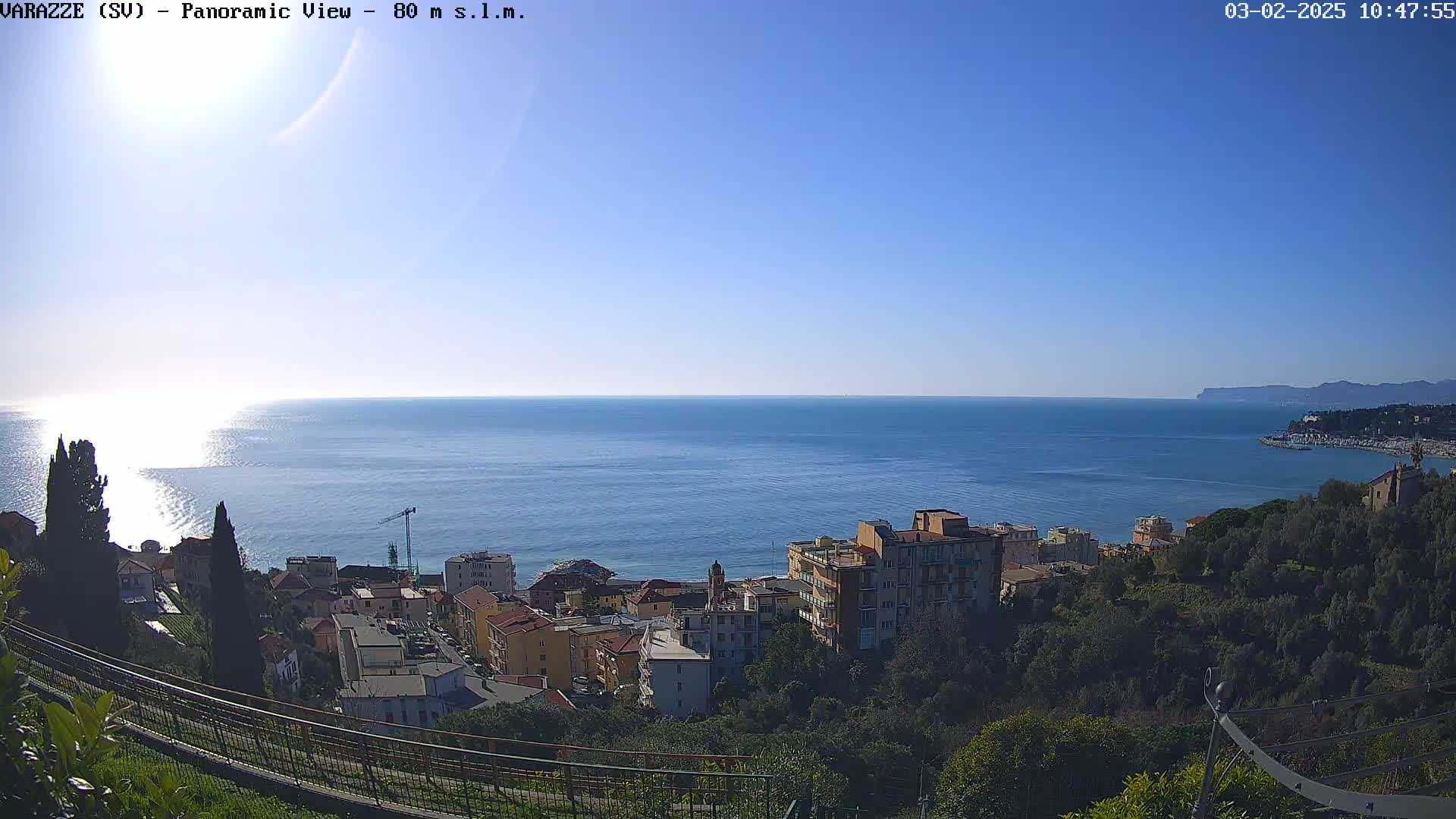 A panoramic view of a coastal town on a clear, sunny day shows a calm sea extending to the horizon, with buildings nestled amongst greenery on a hillside overlooking the water.