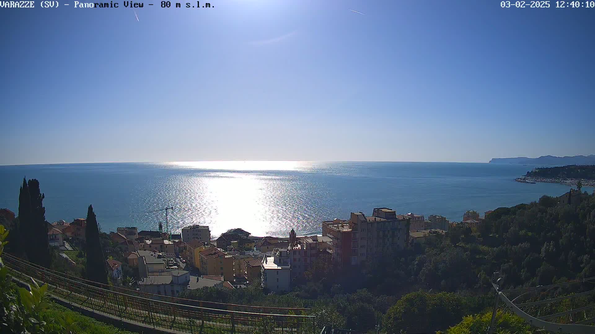 A coastal town nestled on a hillside overlooks a calm, sunlit sea under a clear blue sky.