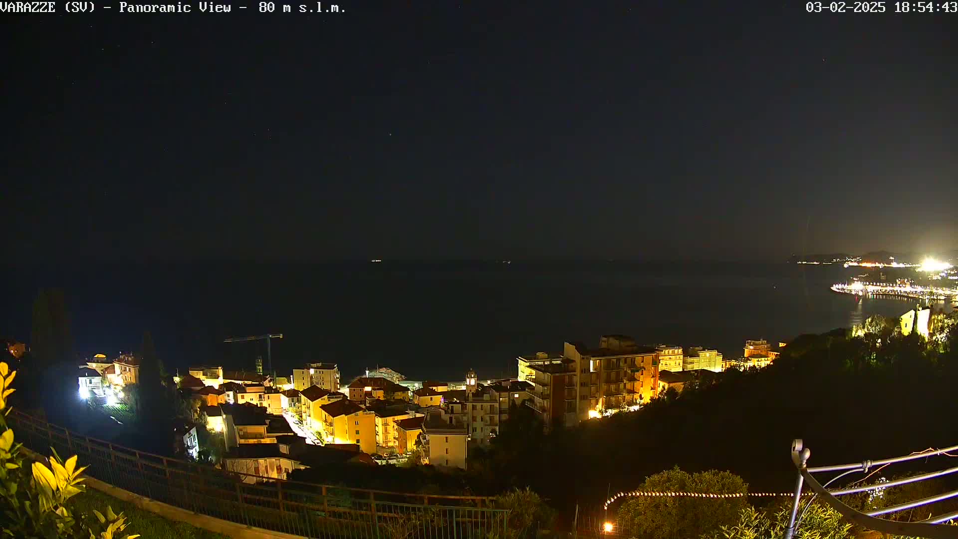A nighttime, panoramic view shows a coastal town illuminated with lights, overlooking a dark body of water under a clear, starry sky.