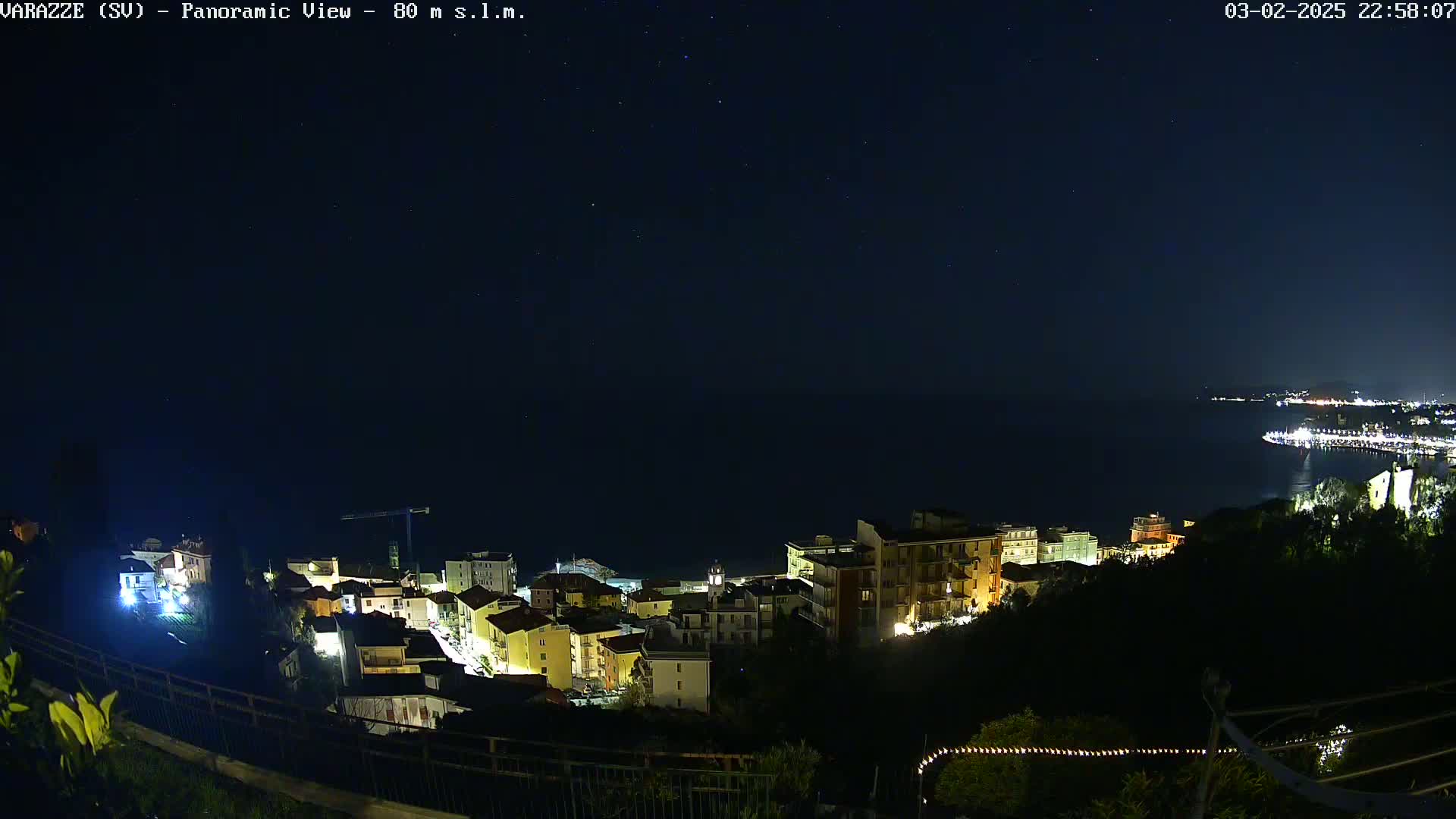 A nighttime, panoramic view shows a coastal town illuminated with lights against a dark sea and starry sky.