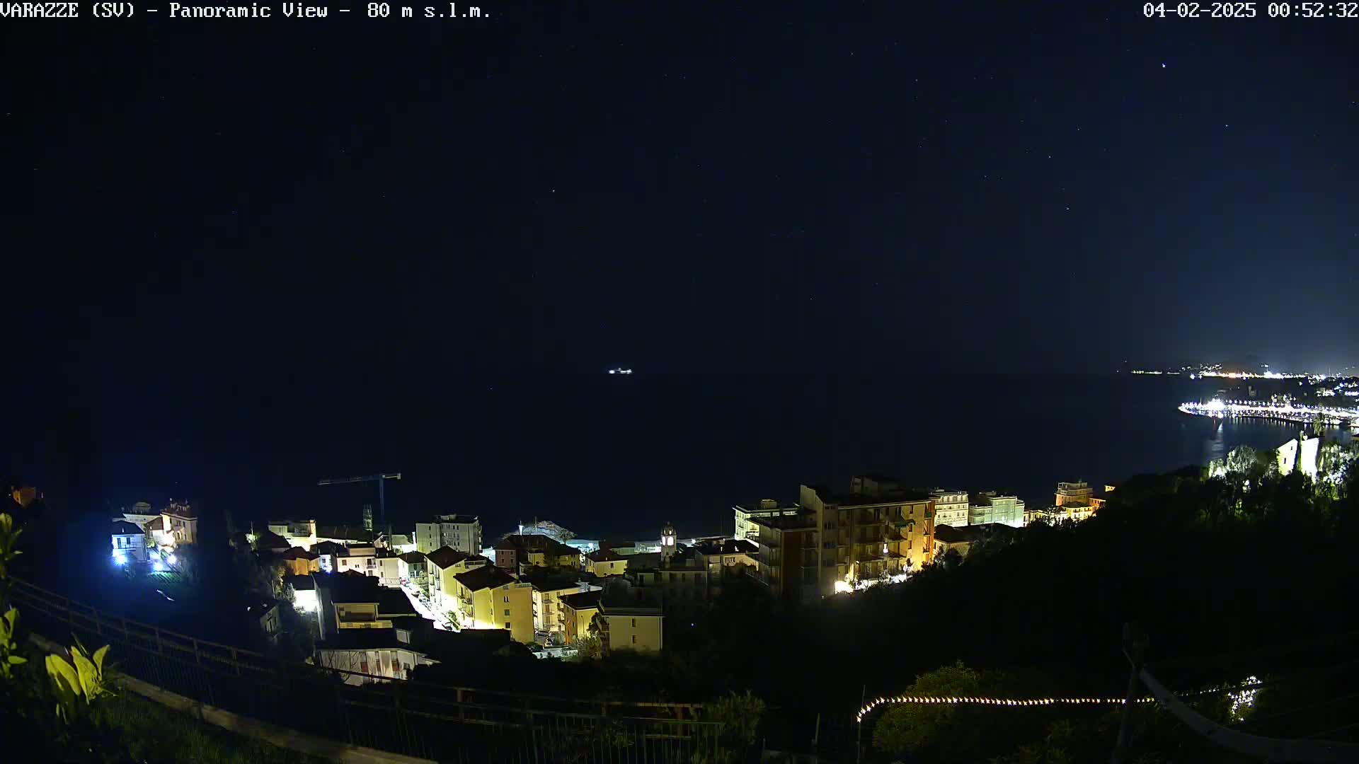 A nighttime panoramic view shows a coastal town illuminated, with a dark sea and starry sky above.