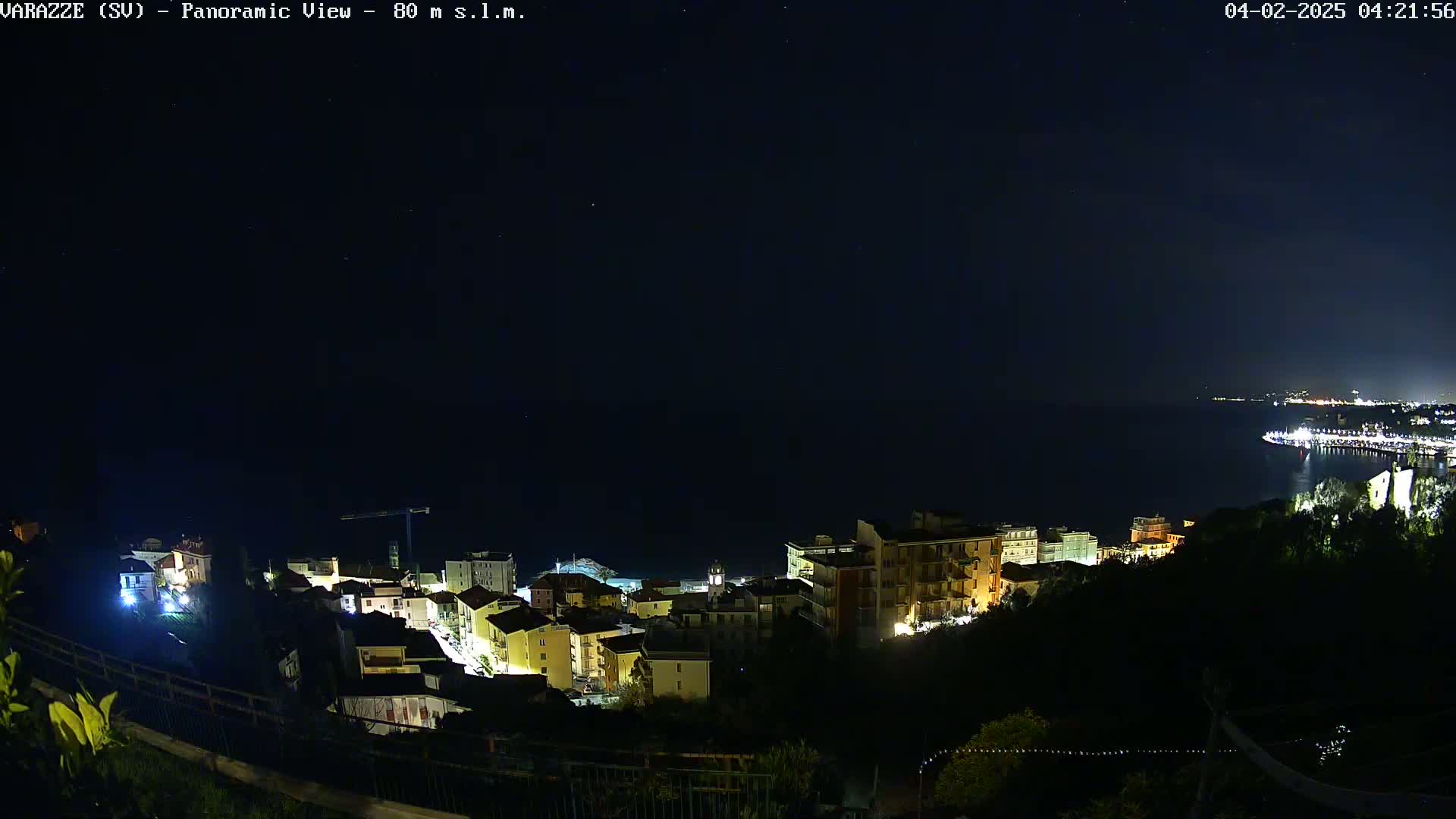 A nighttime, panoramic view shows a coastal town illuminated, with a dark sea and a distant, faintly lit cityscape under a clear, starry sky.