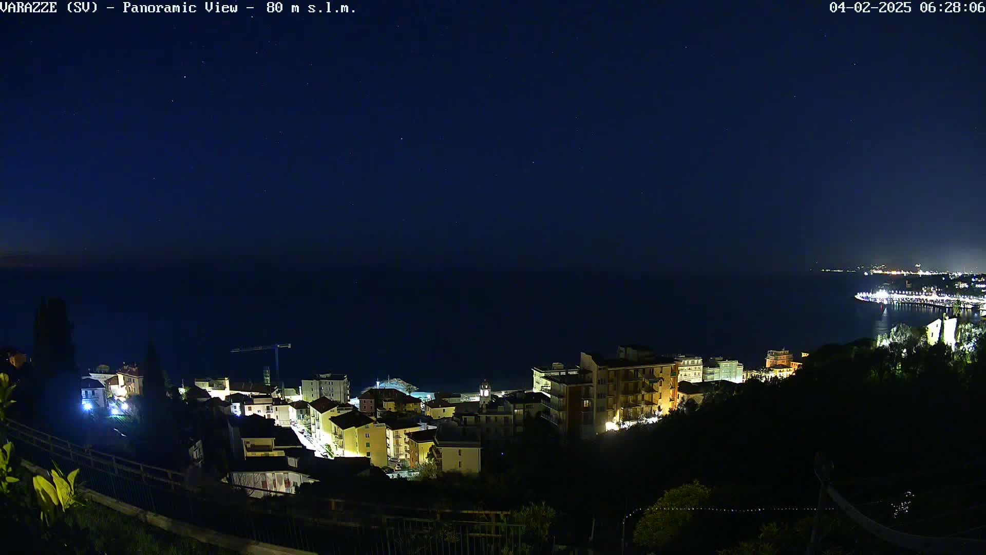 A nighttime panoramic view shows a coastal town with buildings illuminated against a dark blue sea under a clear, starry sky.