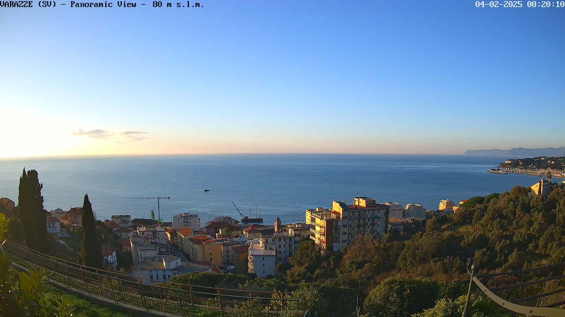 A panoramic view of a coastal town nestled on a hillside overlooking a calm sea under a clear blue sky.