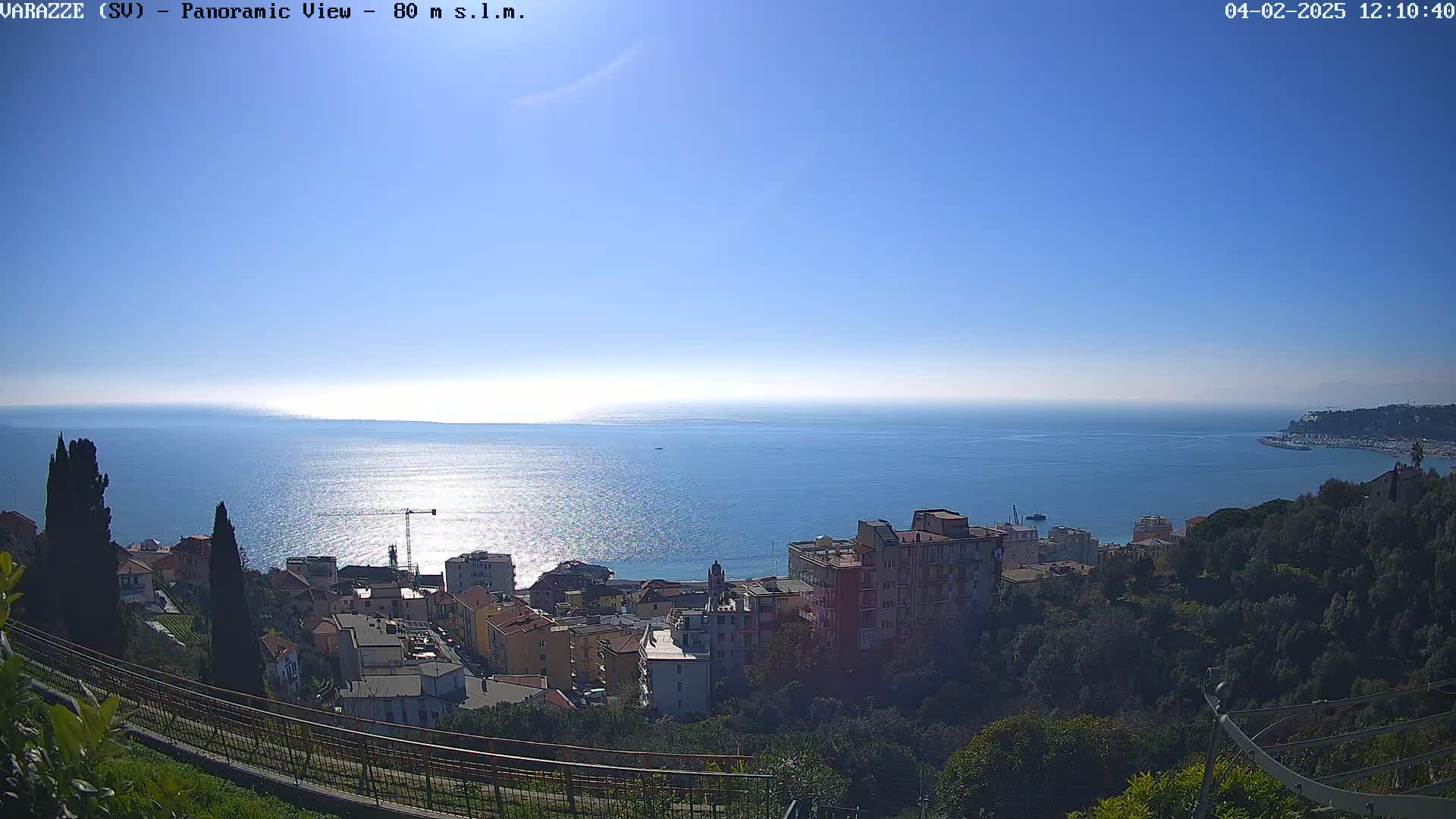 A panoramic view of a coastal town on a sunny day shows a calm blue sea extending to the horizon, with buildings nestled among green hillsides.