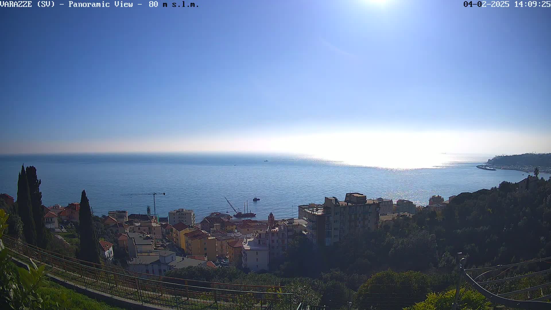 A coastal town nestled on a hillside overlooks a calm, sunlit sea under a clear blue sky.