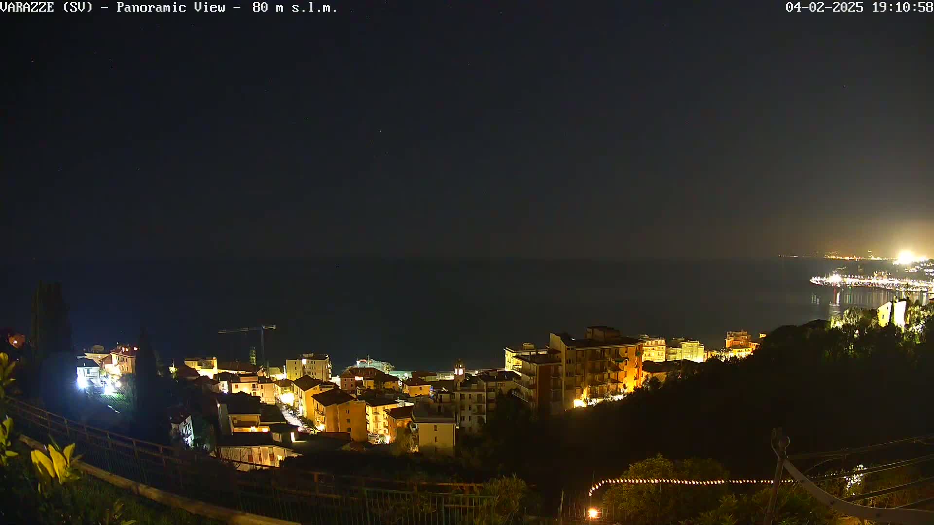 A nighttime panoramic view shows a coastal town with illuminated buildings situated on a hillside overlooking a dark sea under a clear, starry sky.