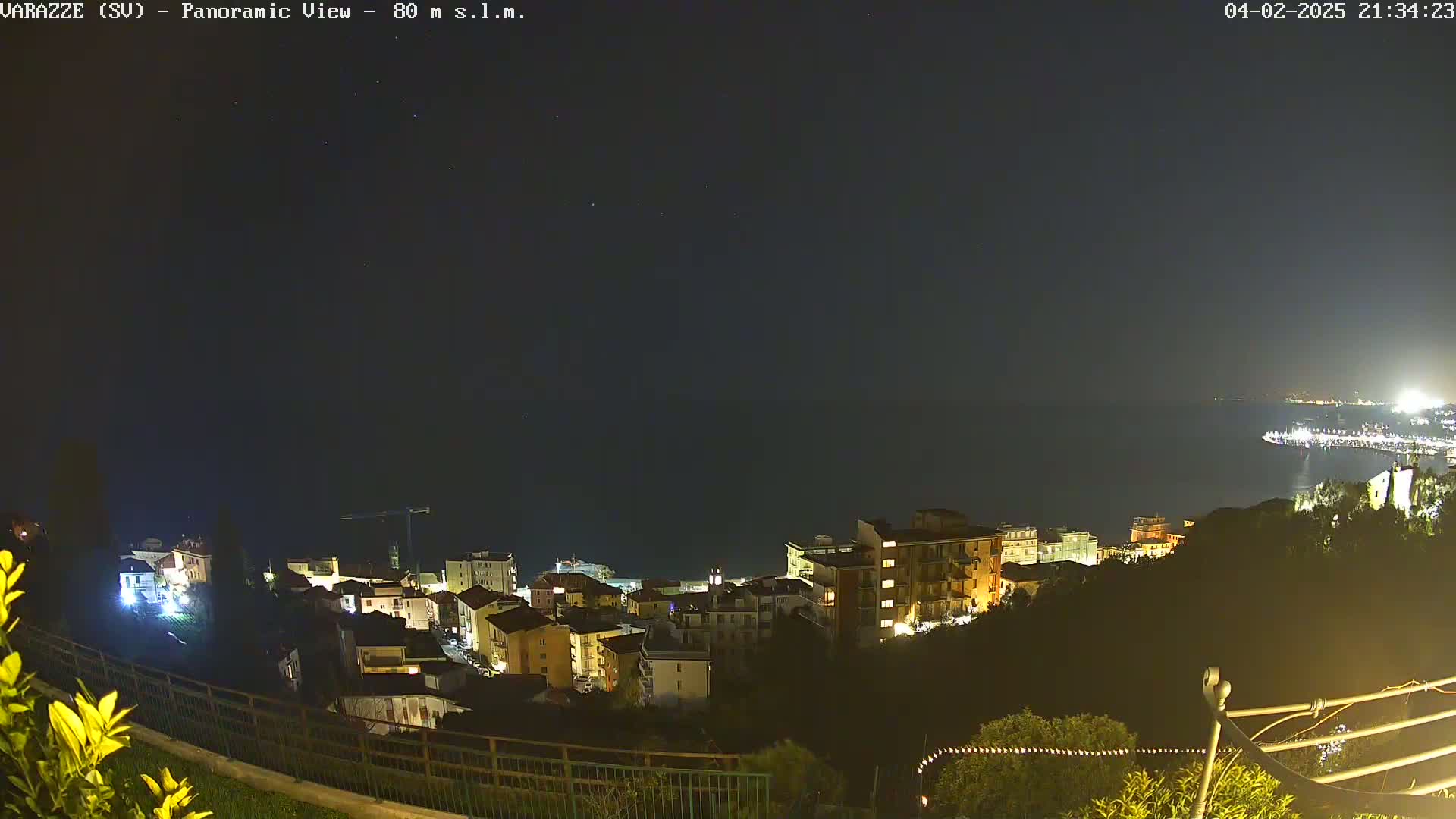 A nighttime, panoramic view shows a coastal town illuminated with lights, overlooking a dark body of water under a clear, starry sky.