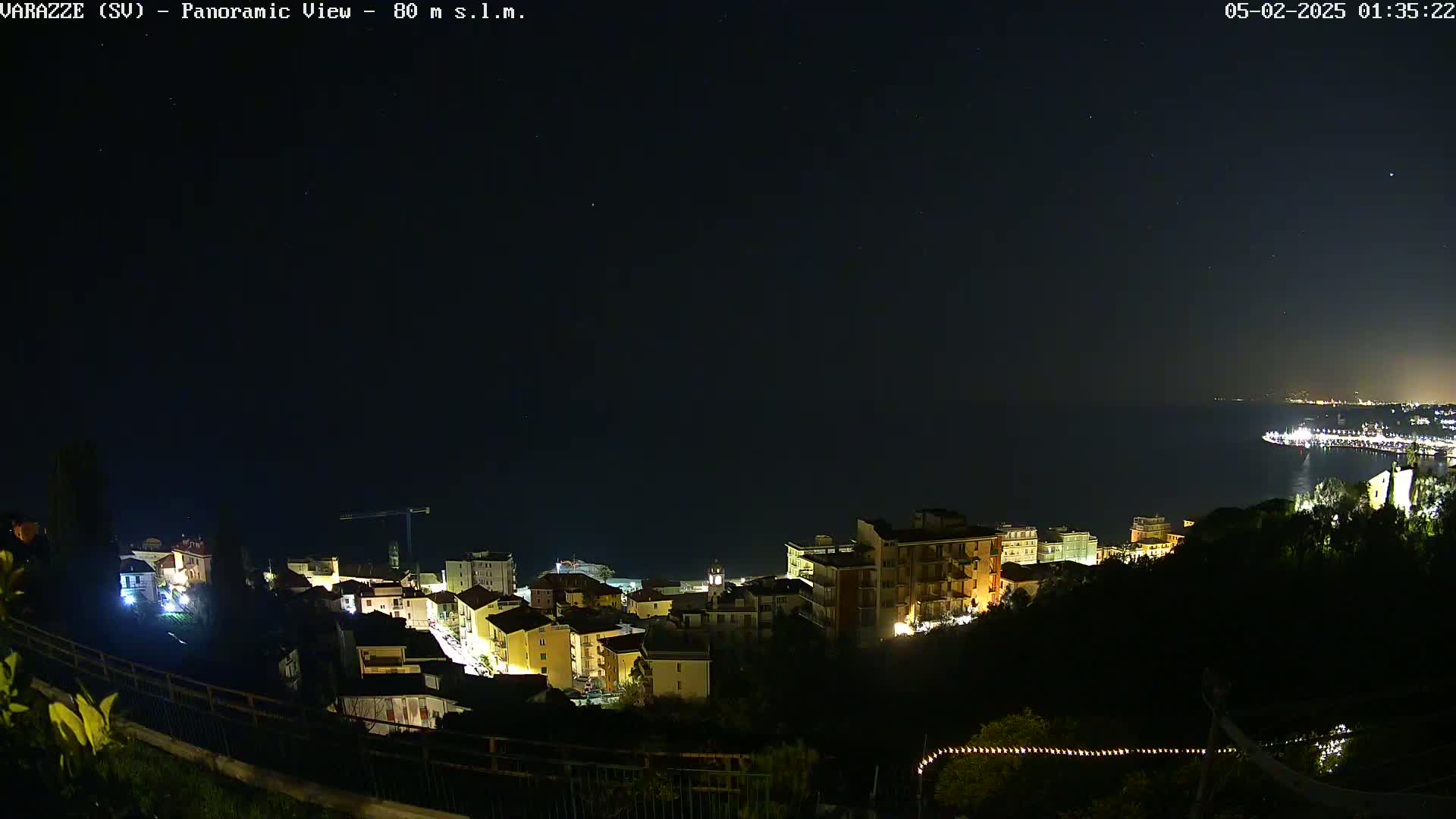 A nighttime panoramic view shows a coastal town with buildings illuminated against a dark sky speckled with stars and a calm sea.