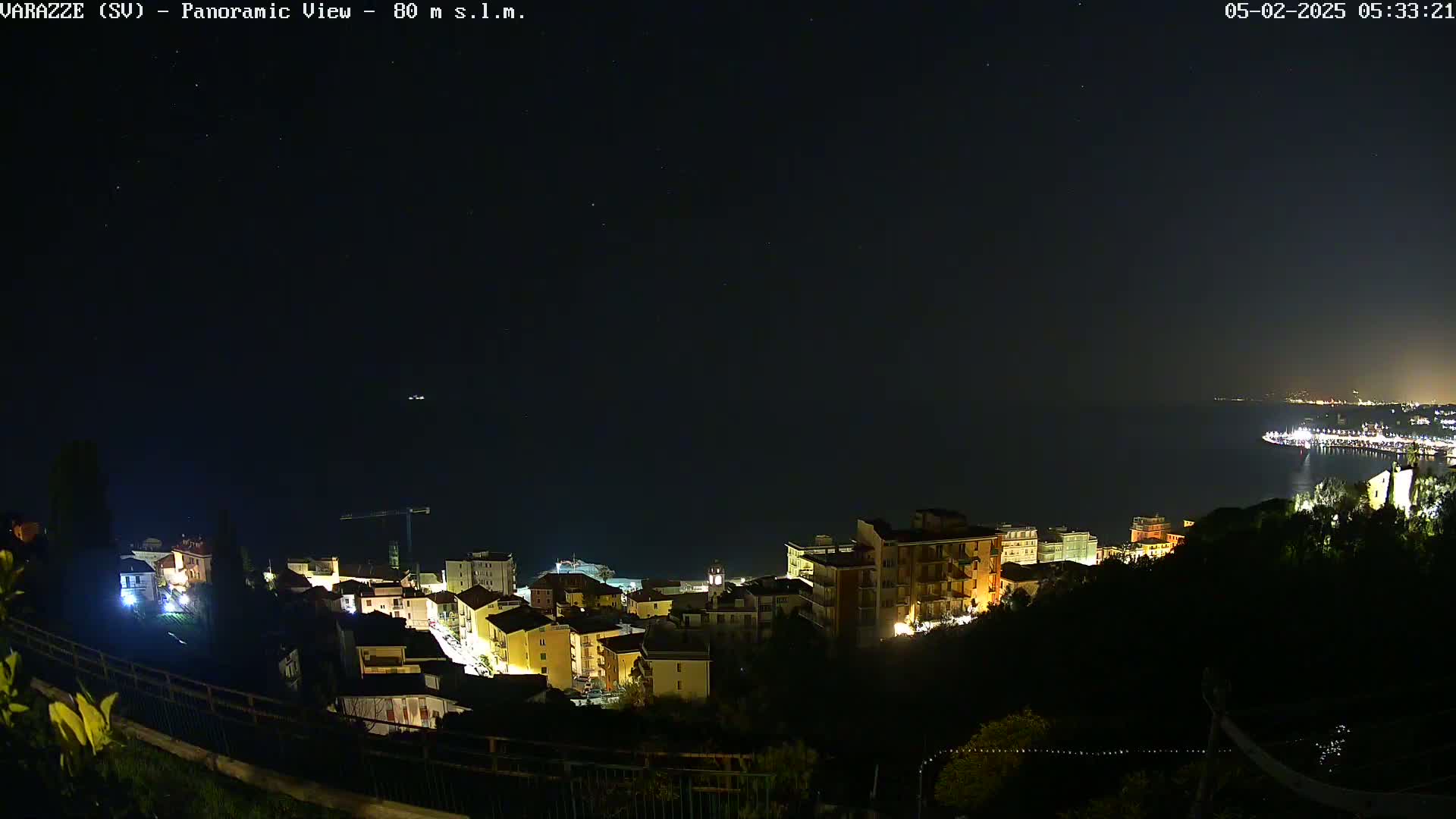 A nighttime panoramic view shows a coastal town illuminated, overlooking a dark body of water under a clear, starry sky.