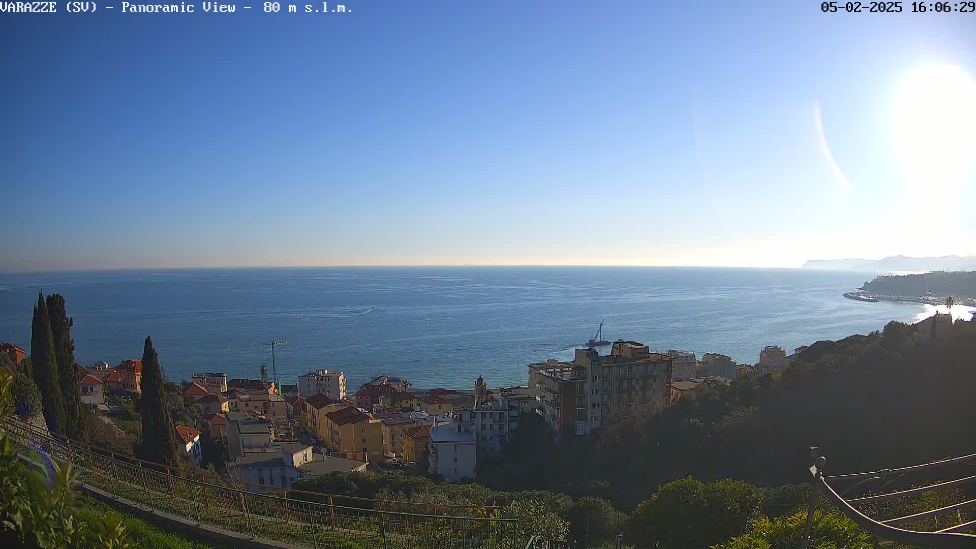 A coastal town with buildings nestled on a hillside overlooking a calm, blue sea under a clear, sunny sky.