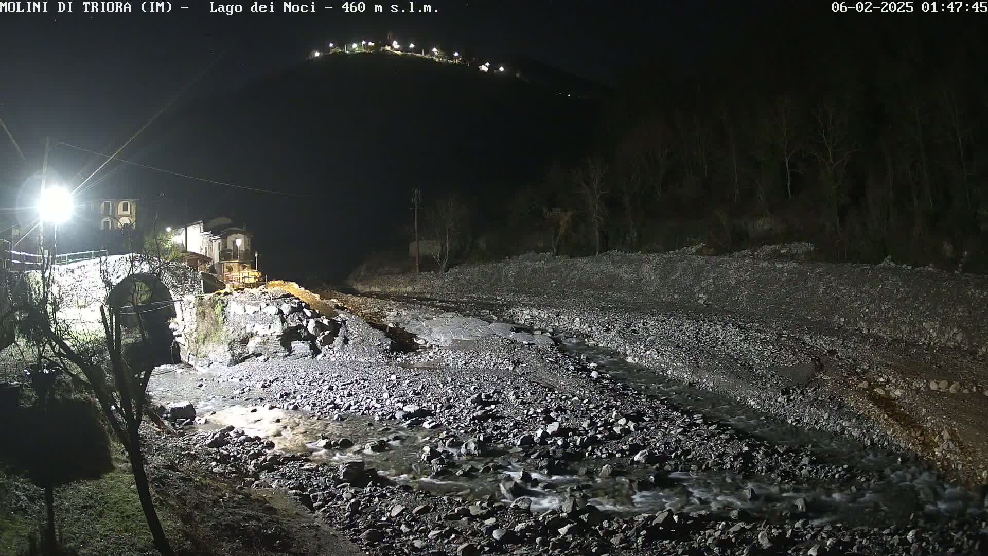 At night, a rocky stream flows past a stone bridge and small village under artificial light.
