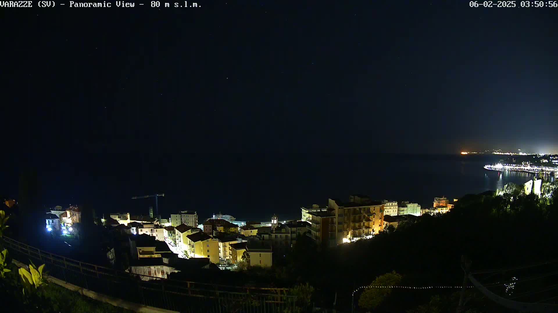A nighttime panoramic view shows a coastal town illuminated, with a dark, clear sky above and a calm body of water in the distance.