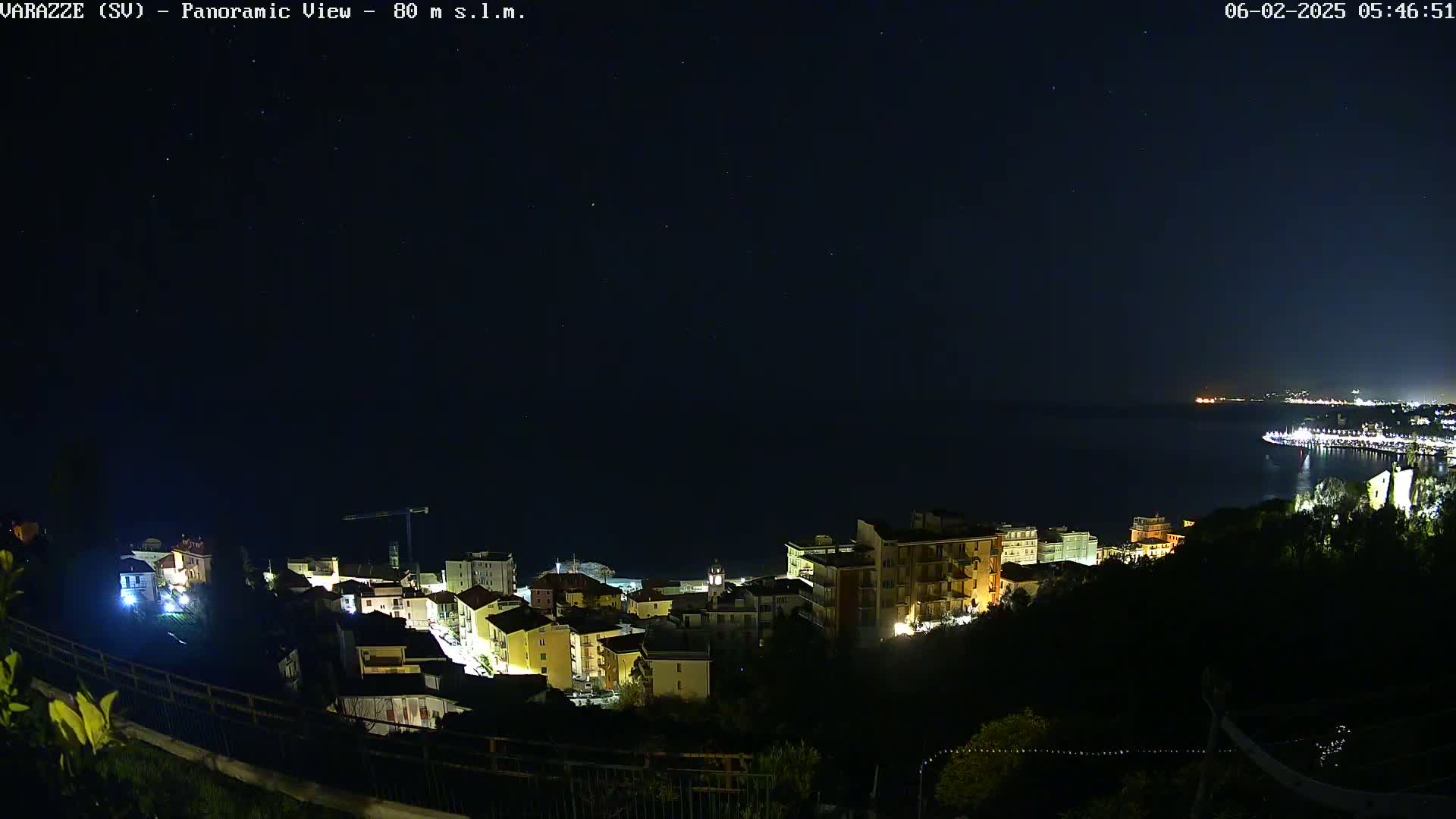 A nighttime panoramic view shows a coastal town illuminated, with a dark sea and a starry sky above.