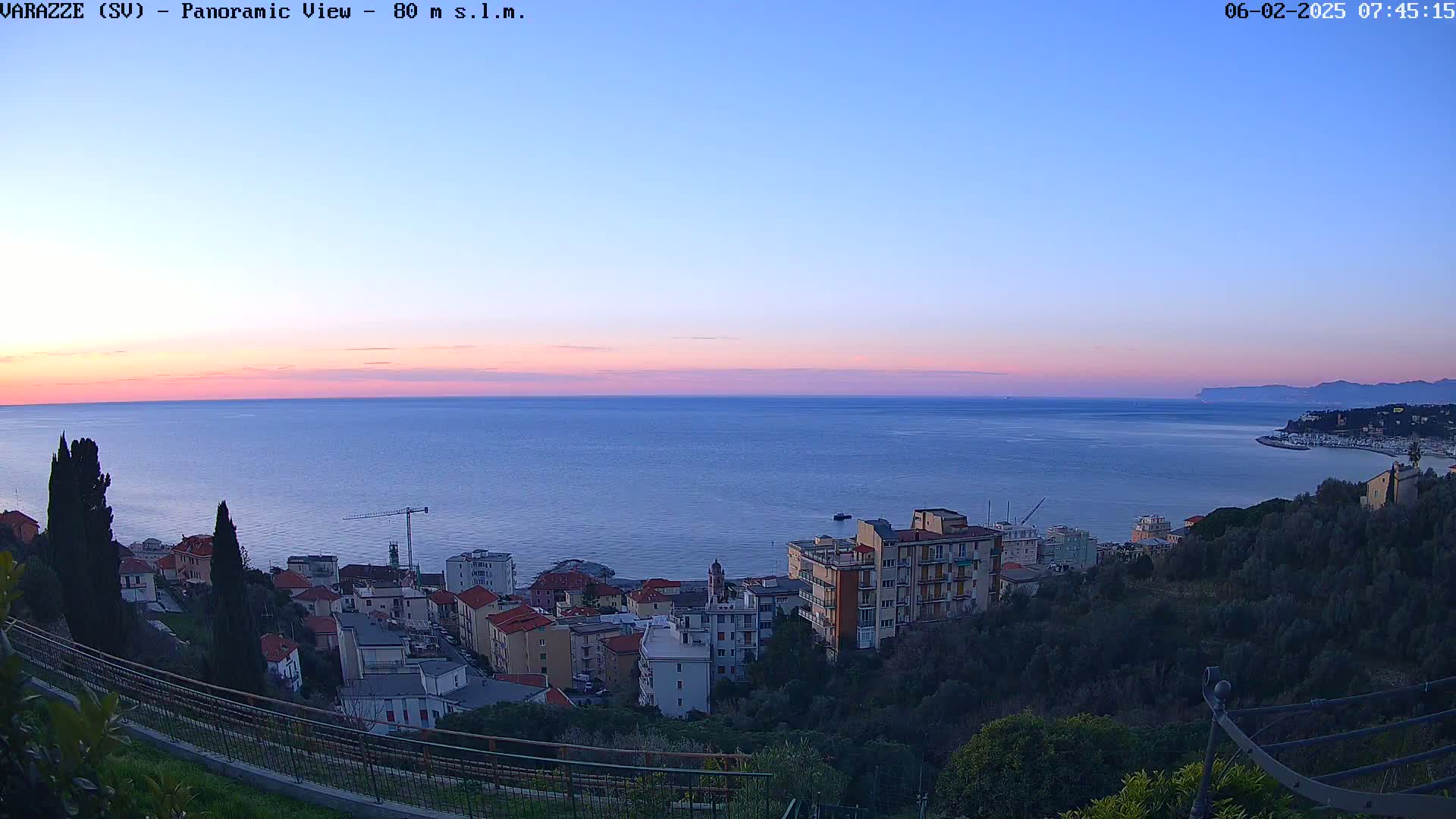 A panoramic view at sunset shows a coastal town nestled on a hillside overlooking a calm sea, under a clear, twilight sky.