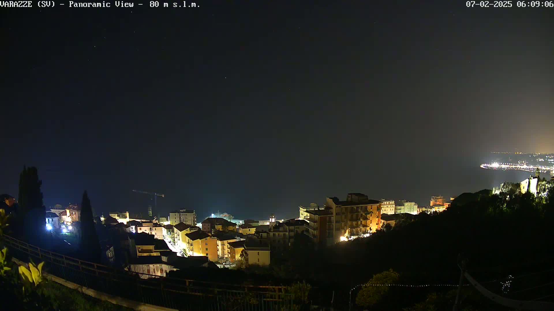 A nighttime, panoramic view of a coastal town, illuminated by streetlights, with a dark sky and calm sea visible in the background.