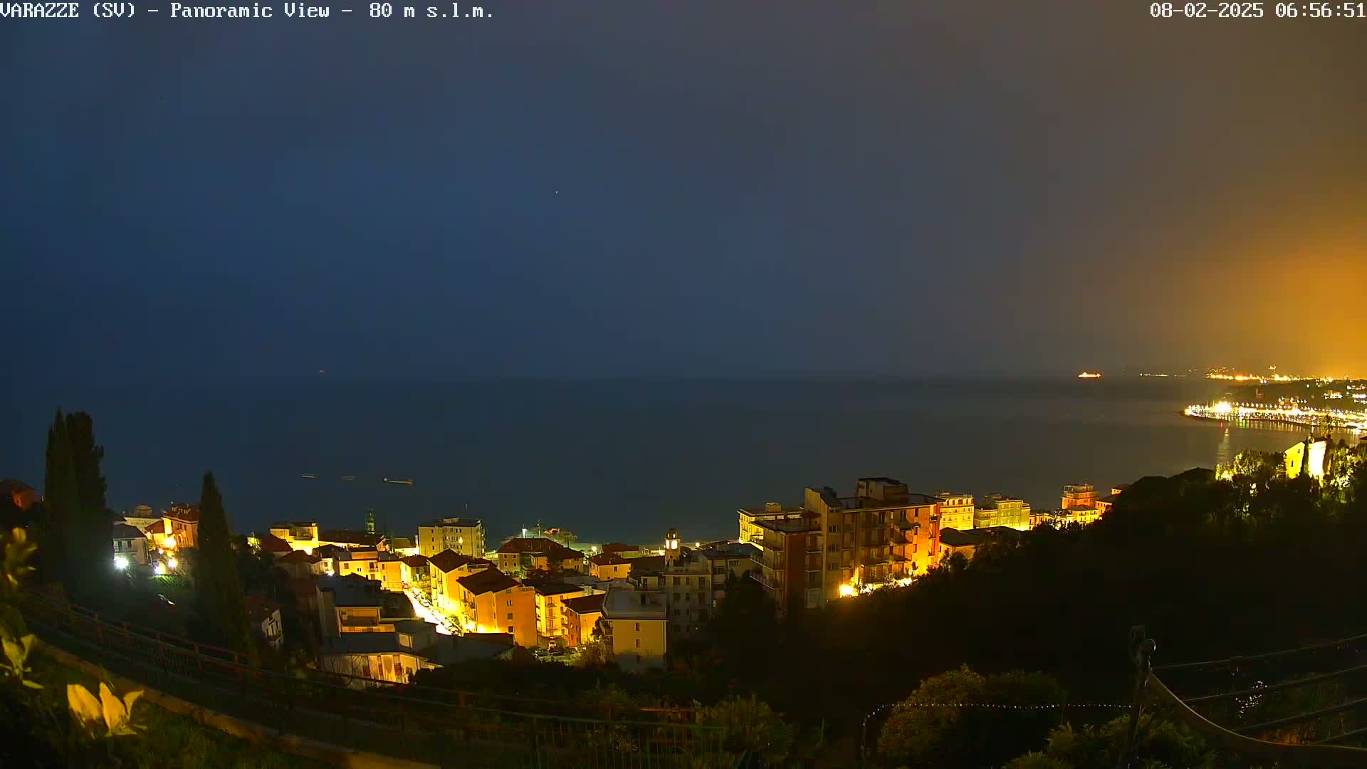 A nighttime panoramic view shows a coastal town illuminated, nestled on a hillside overlooking a dark, calm sea under a twilight sky.