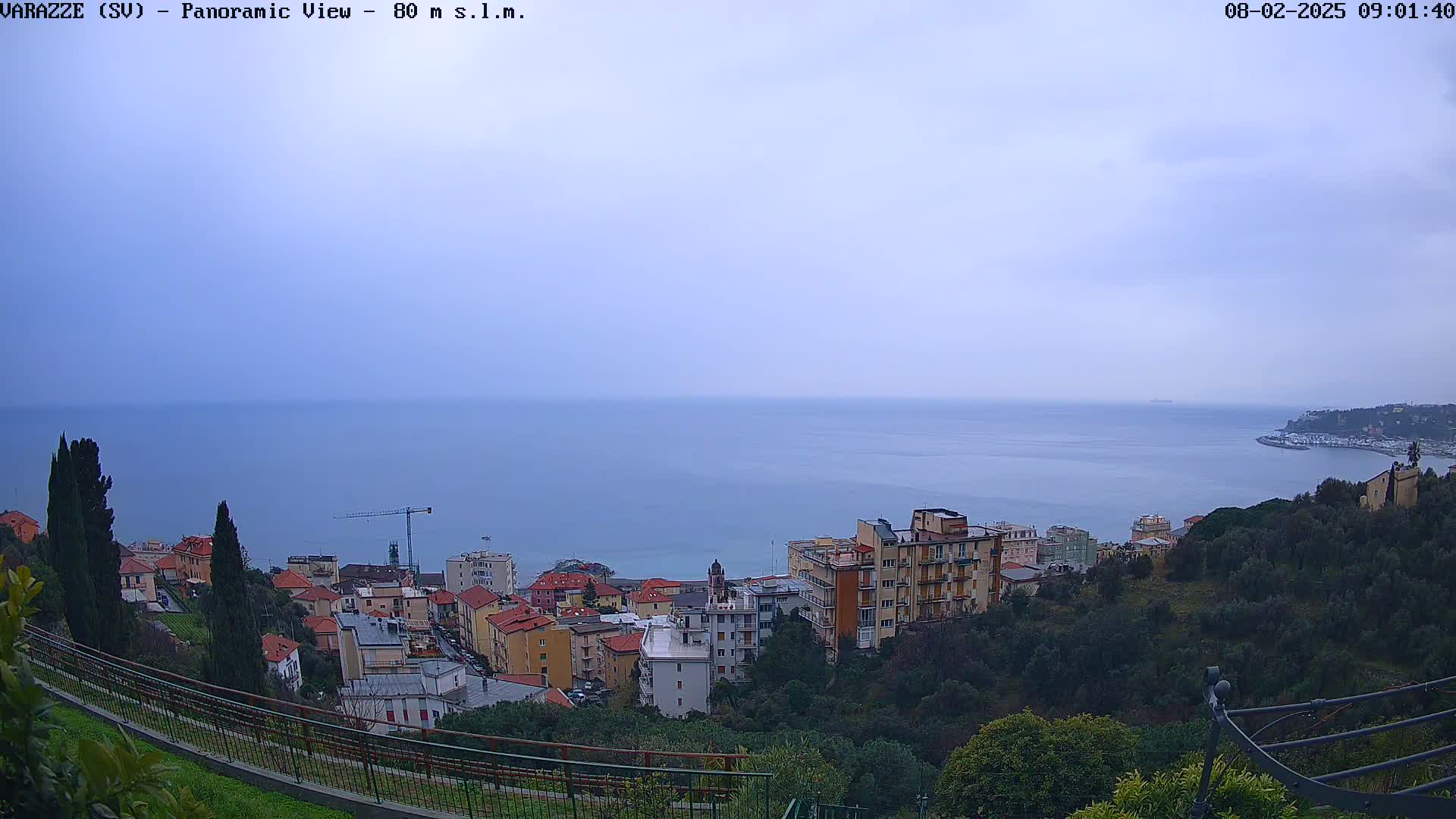 A coastal town with multi-story buildings is nestled on a hillside overlooking a calm sea under an overcast sky.