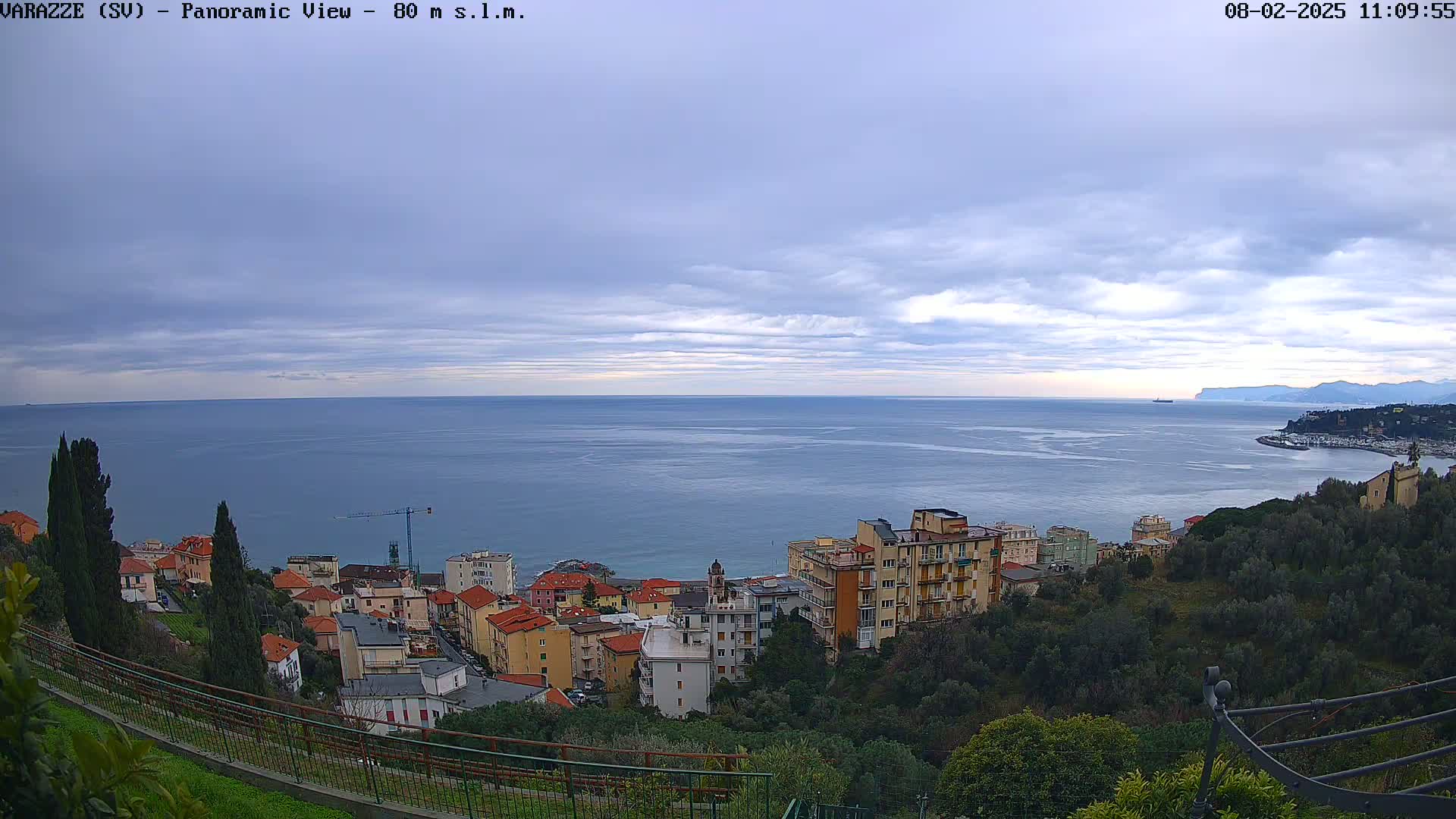 A panoramic view of a coastal town under a cloudy sky shows a calm sea extending to distant mountains.