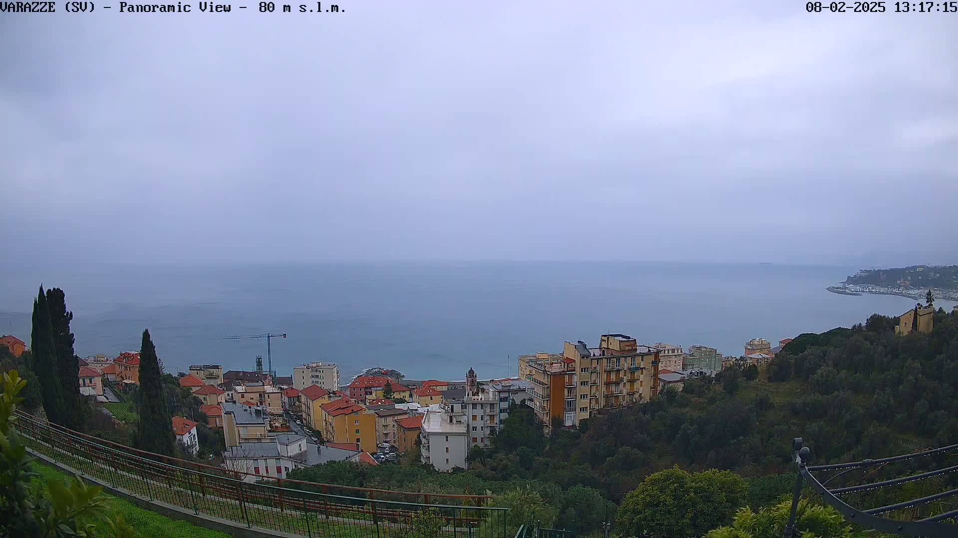 A panoramic view of a coastal town on a cloudy day shows a calm sea and multi-story buildings nestled among green hills.