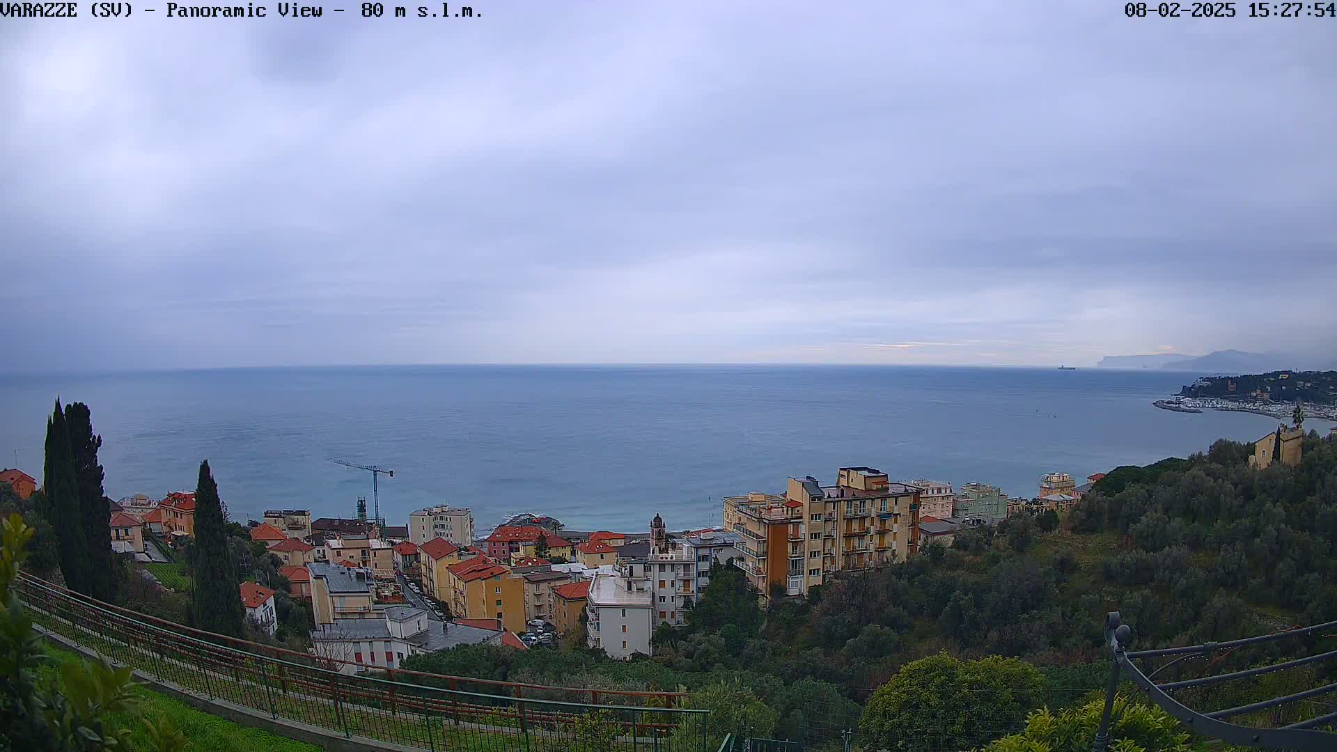 A panoramic view shows a coastal town nestled on a hillside overlooking a calm sea under a cloudy sky.