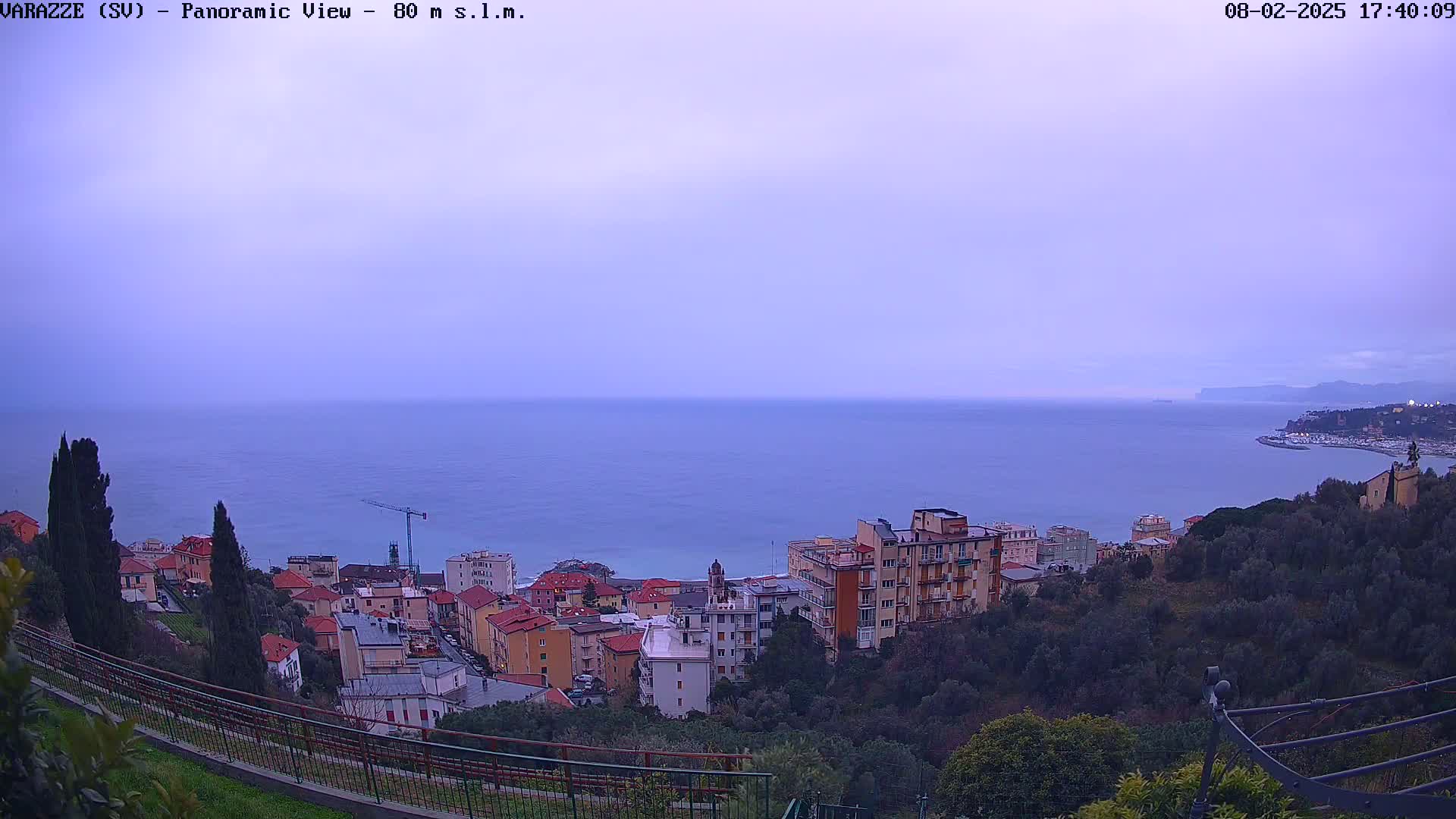 A panoramic view of a coastal town under a dusky sky shows a calm sea and buildings nestled into a hillside.