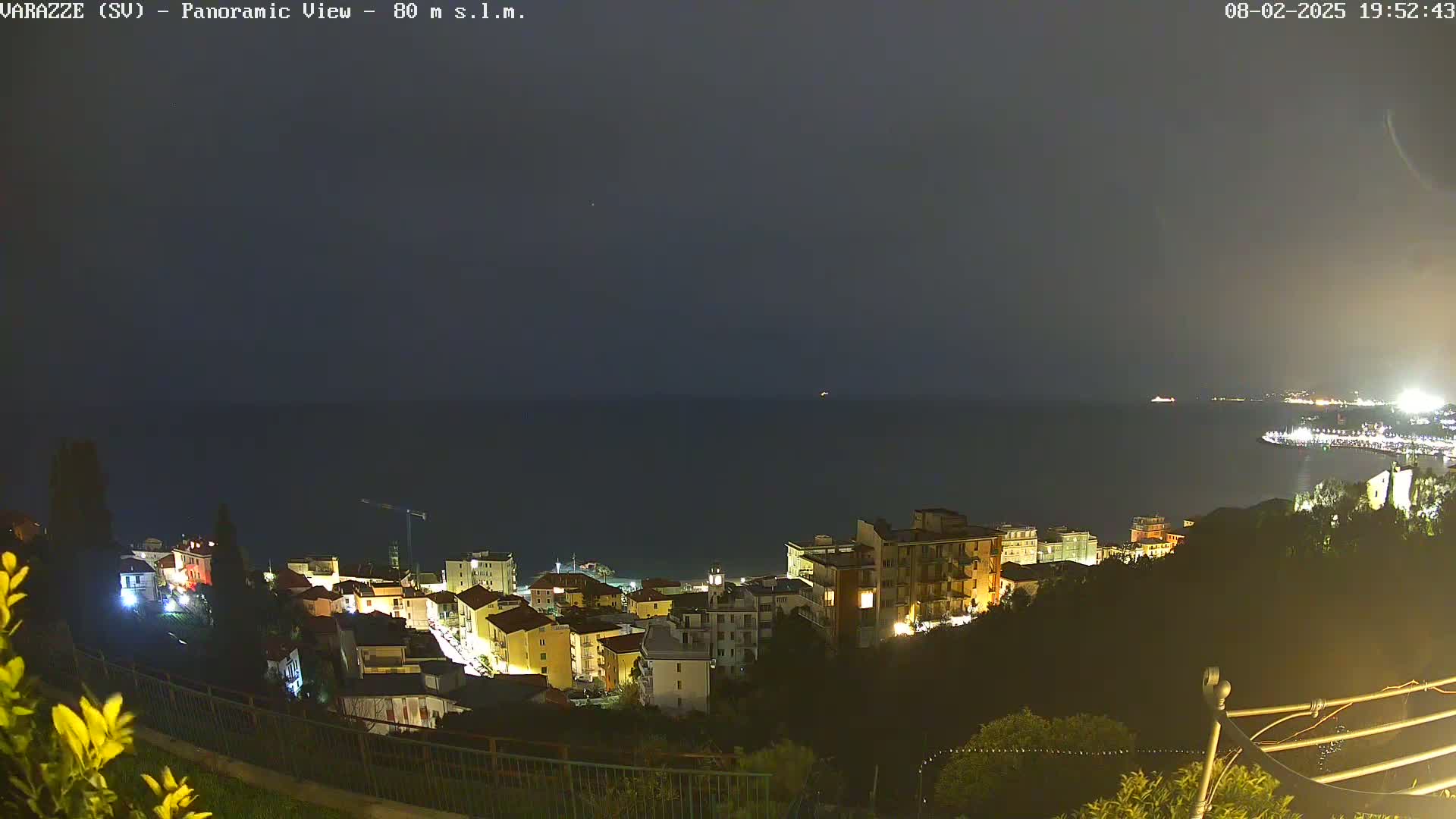 A nighttime, panoramic view shows a coastal town illuminated with lights, overlooking a dark sea under a mostly clear sky.
