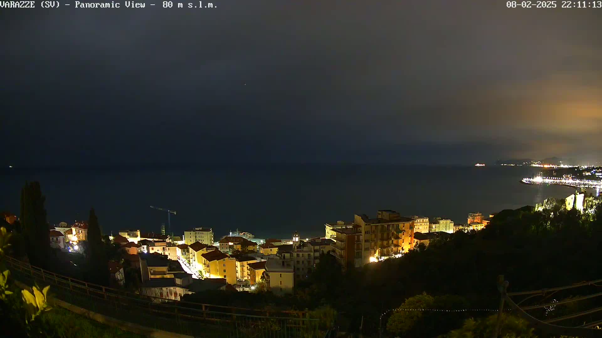 A nighttime panoramic view shows a coastal town illuminated against a dark, overcast sky and a calm, dark sea.