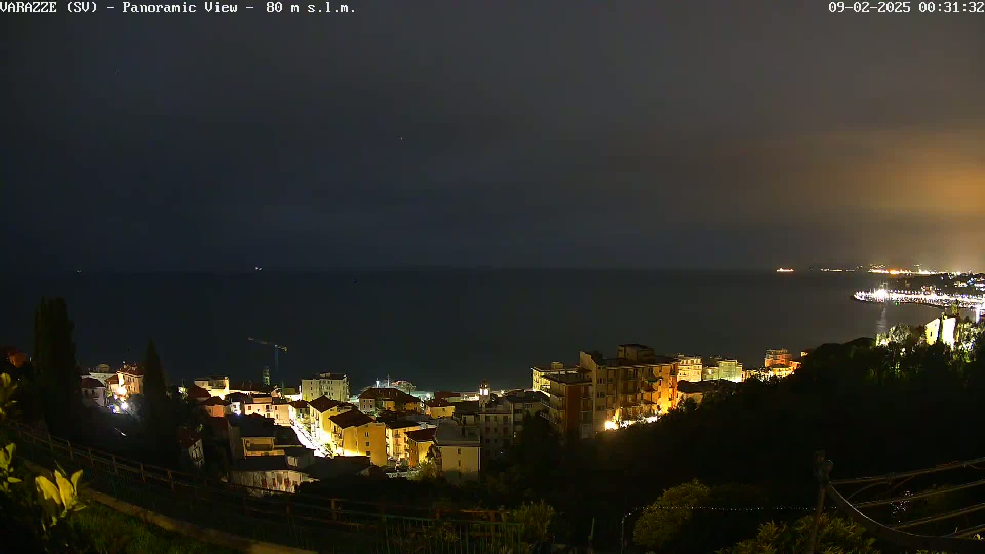 A nighttime panoramic view of a coastal town illuminated by streetlights, overlooking a dark sea under a cloudy sky.