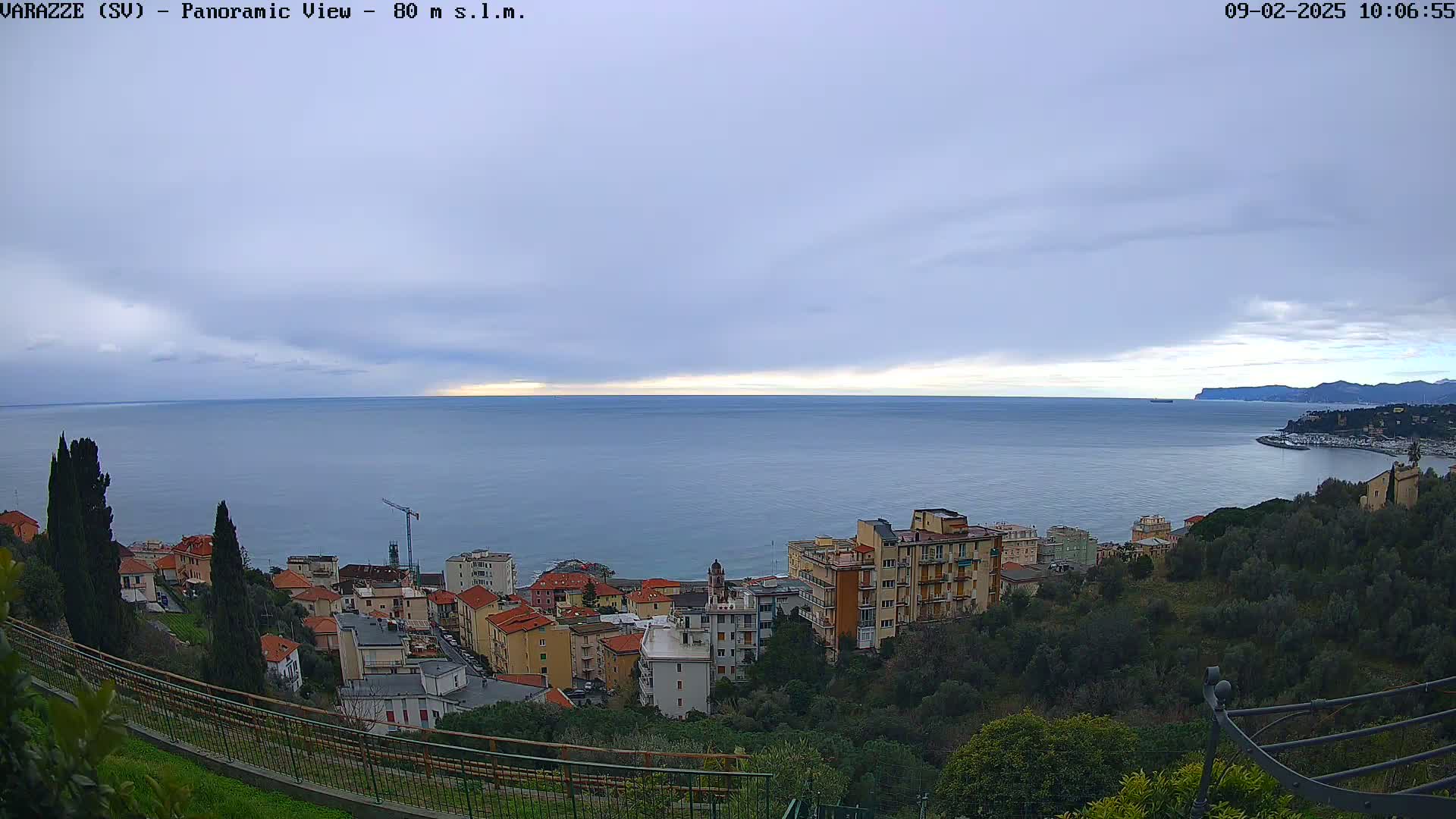 A panoramic view shows a calm sea under an overcast sky, with a coastal town nestled on a hillside overlooking the water.