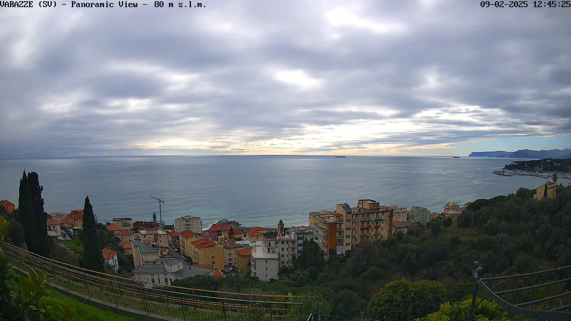 A coastal town with multi-story buildings is nestled on a hillside overlooking a calm sea under a mostly cloudy sky.