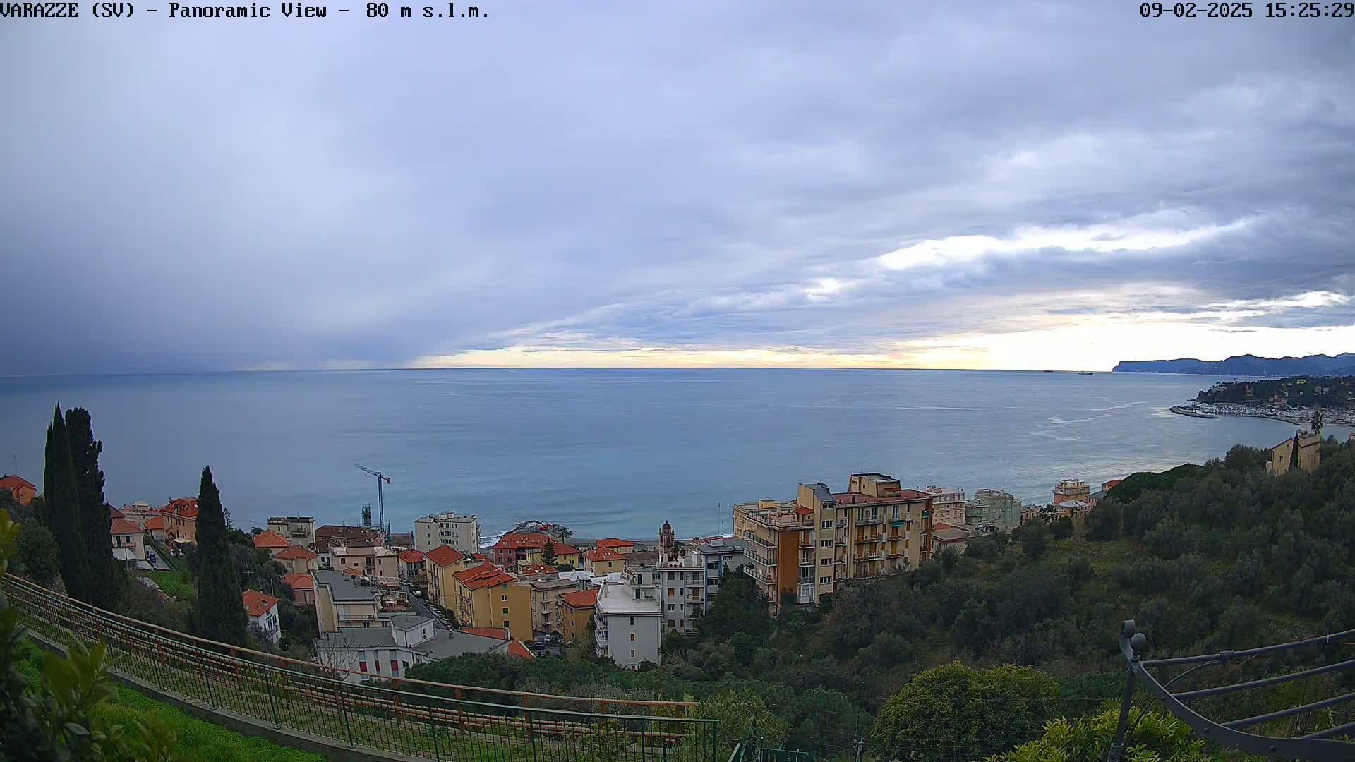 A coastal town nestled on a hillside overlooks a calm sea under a mostly cloudy sky.