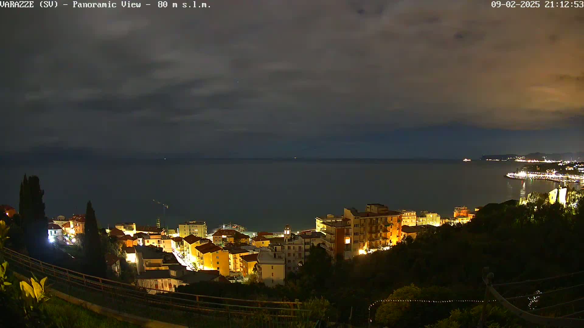A nighttime, slightly cloudy view overlooks a coastal town illuminated by streetlights, with a dark body of water extending to a distant, faintly lit shoreline.