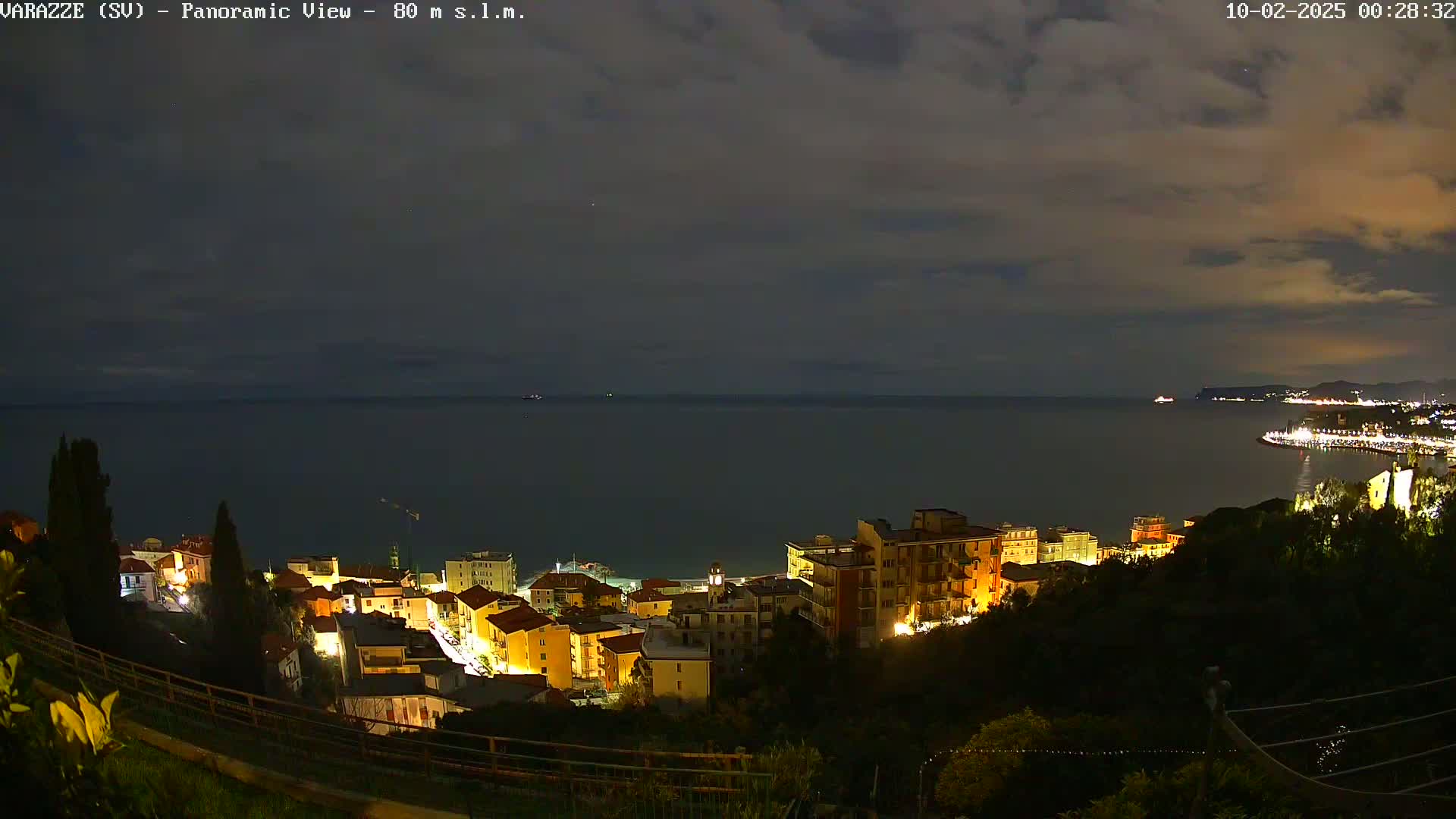 A nighttime, panoramic view of a coastal town with illuminated buildings overlooking a dark sea under a partly cloudy sky.