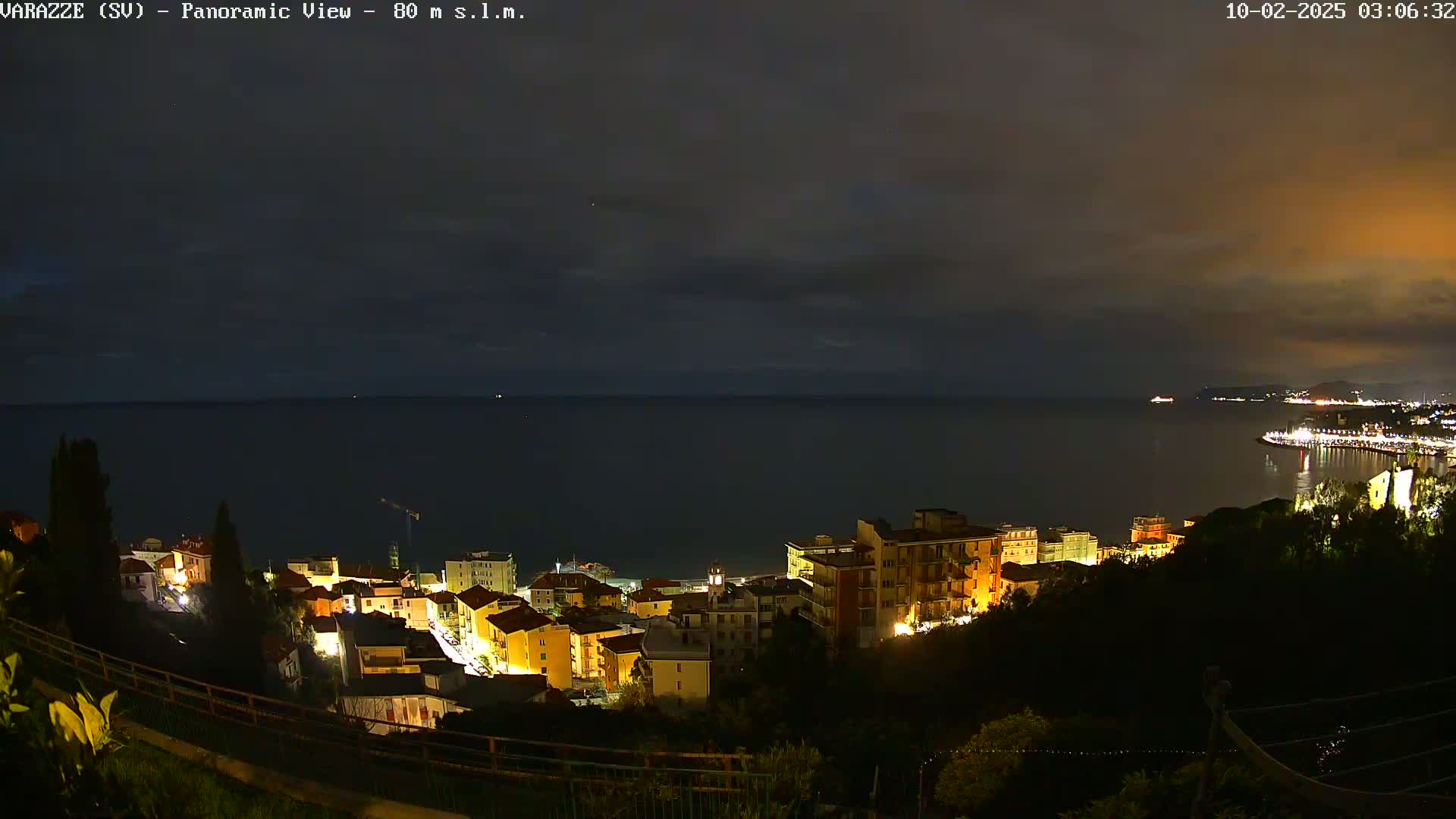 A nighttime panoramic view shows a coastal town illuminated with lights, overlooking a dark sea under a mostly cloudy sky.