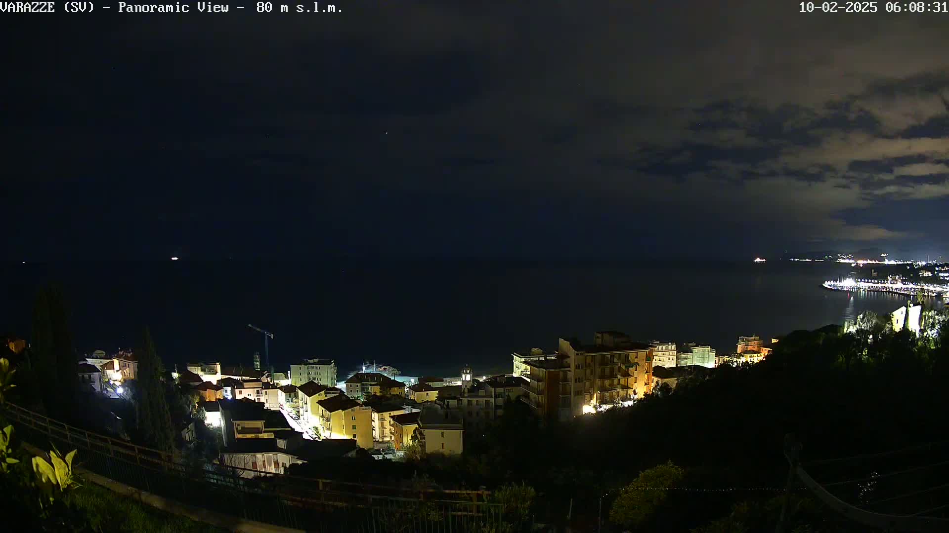 A nighttime panoramic view shows a coastal town illuminated against a dark, cloudy sky over a calm body of water.