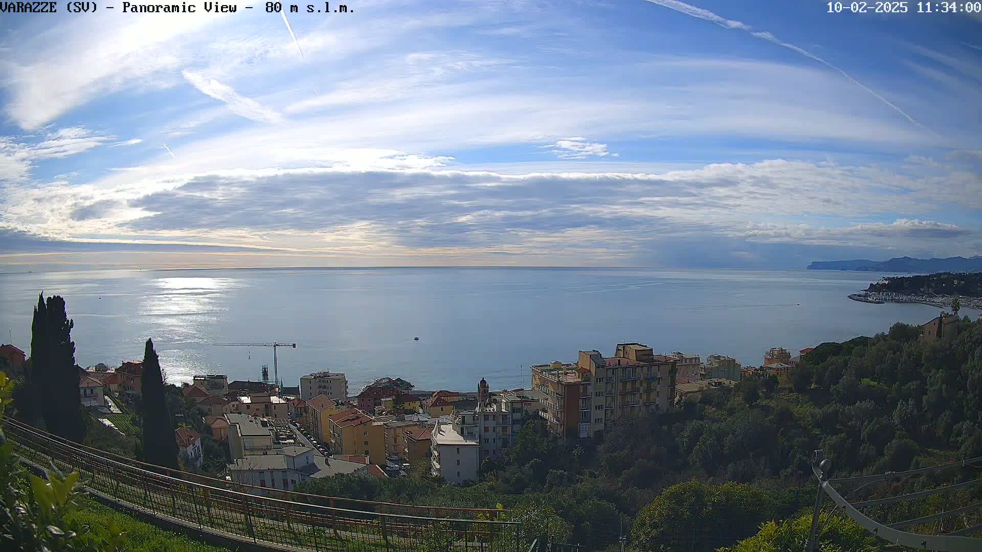A panoramic view shows a partly cloudy sky over a calm sea, with a coastal town nestled below, partially obscured by trees.