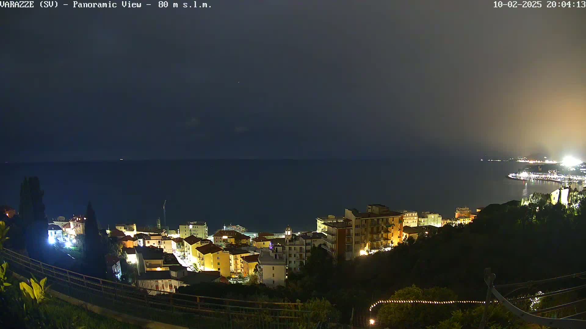 A nighttime panoramic view of a coastal town illuminated, overlooking a dark sea under a mostly clear, dusky sky.