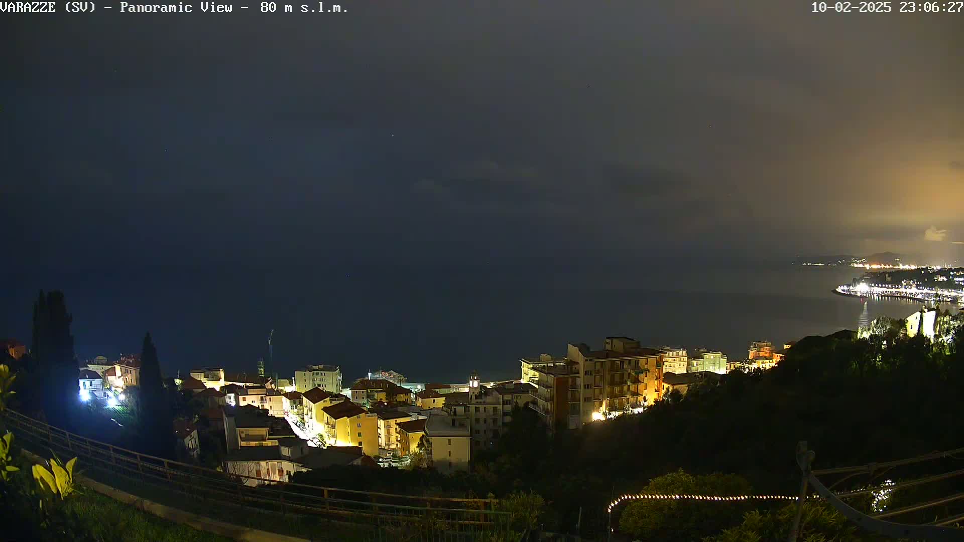 A nighttime, panoramic view of a coastal town illuminated by lights, overlooking a dark sea under a cloudy sky.