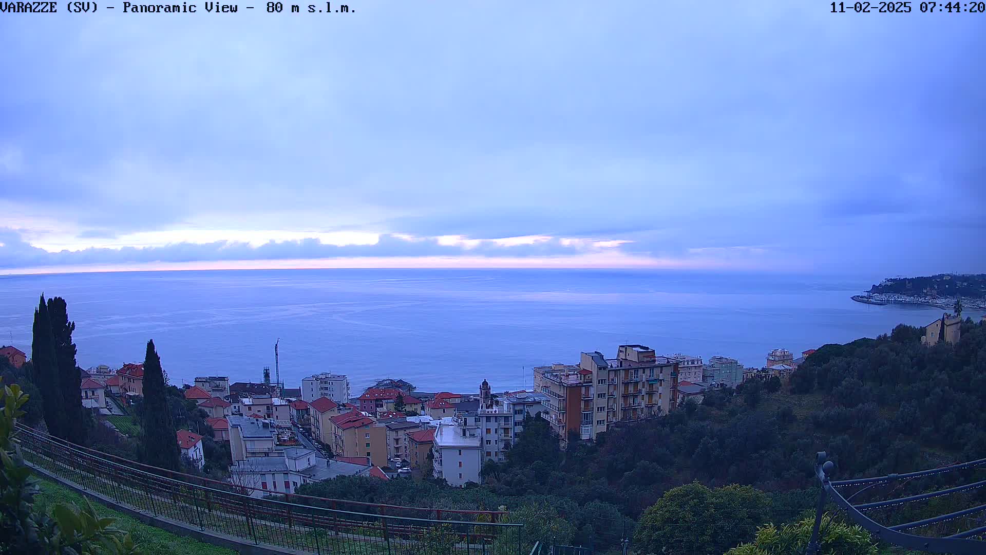 A panoramic view of a coastal town nestled on a hillside overlooking a calm sea under a mostly cloudy, pale sky.
