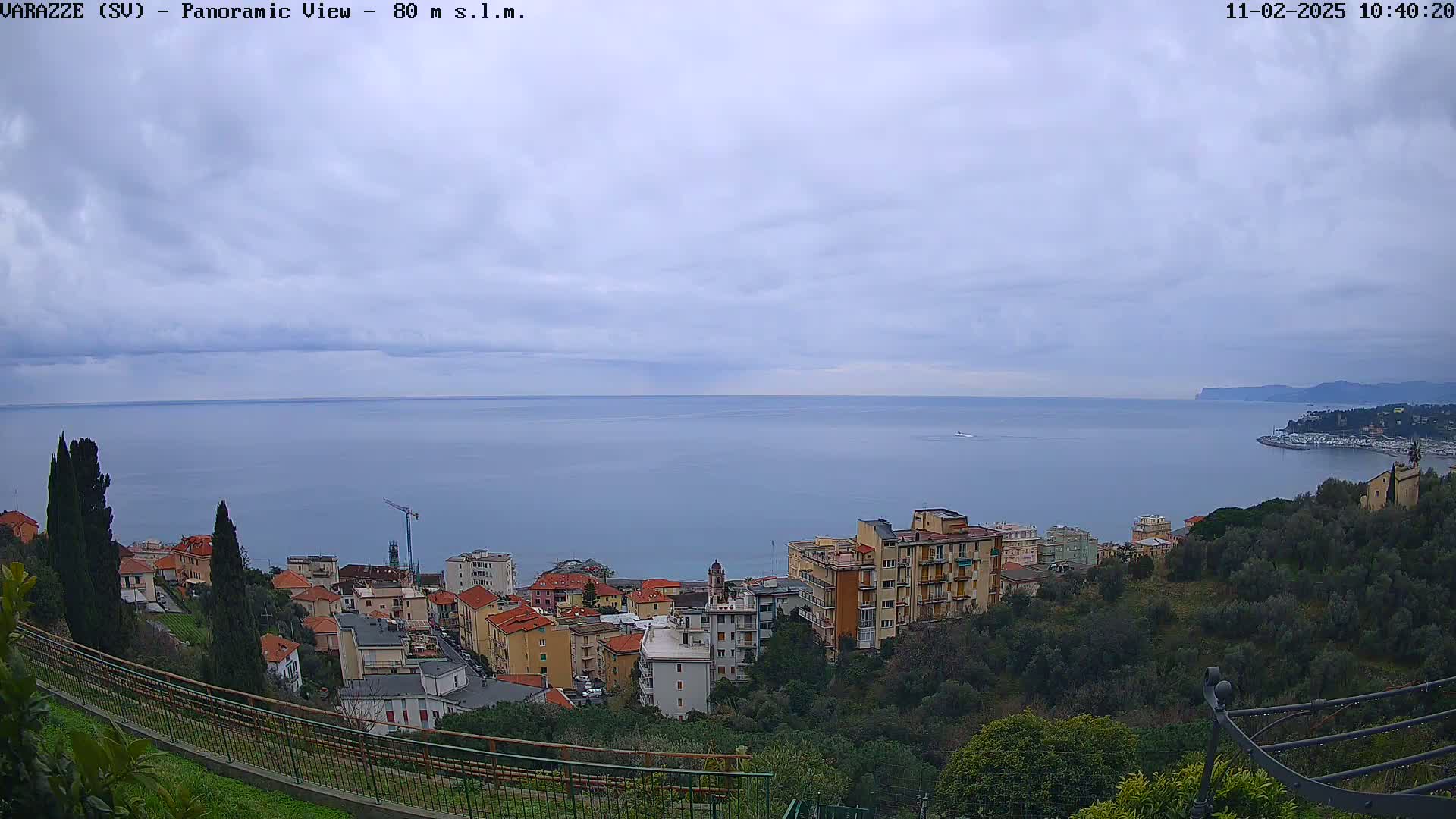A panoramic view shows a coastline town nestled on a hillside overlooking a calm sea under a cloudy sky.