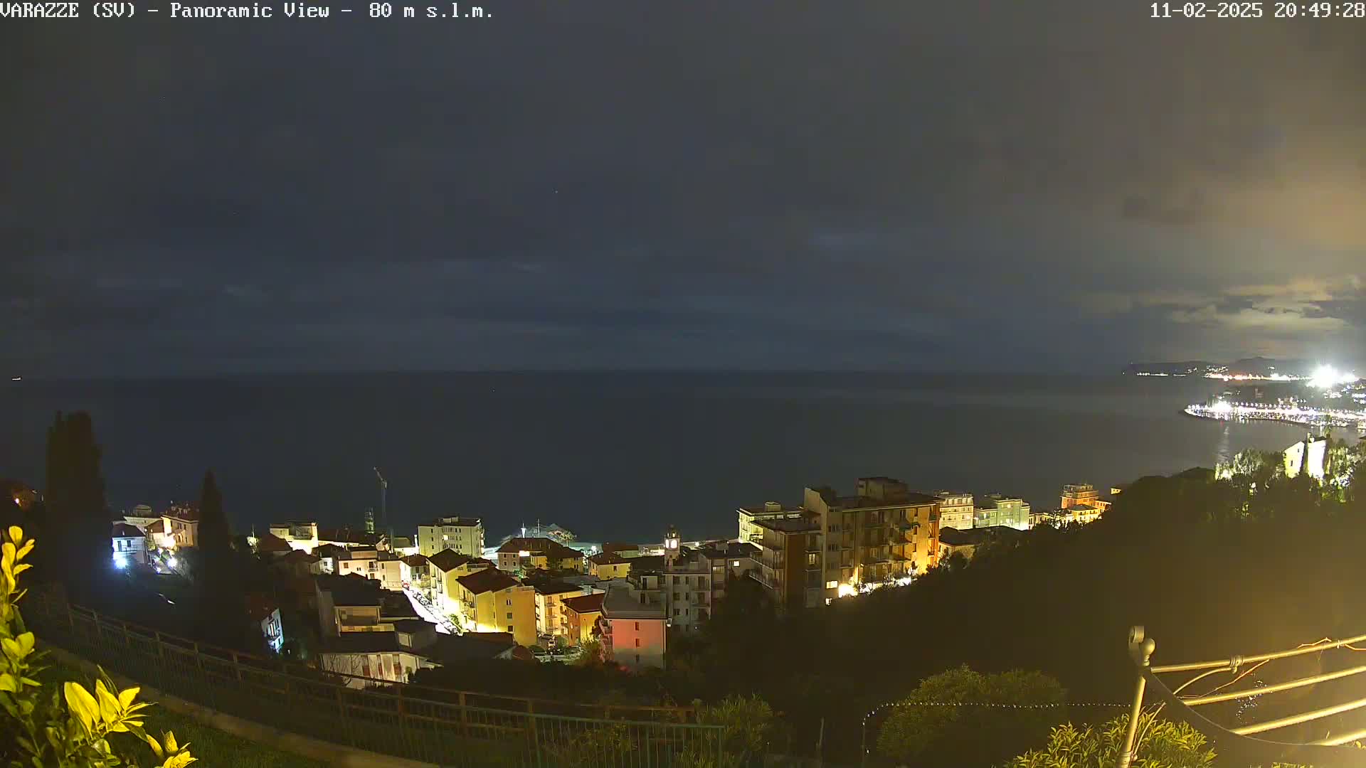 A nighttime panoramic view of a coastal town with its lights illuminated against a dark, calm sea under a cloudy sky.