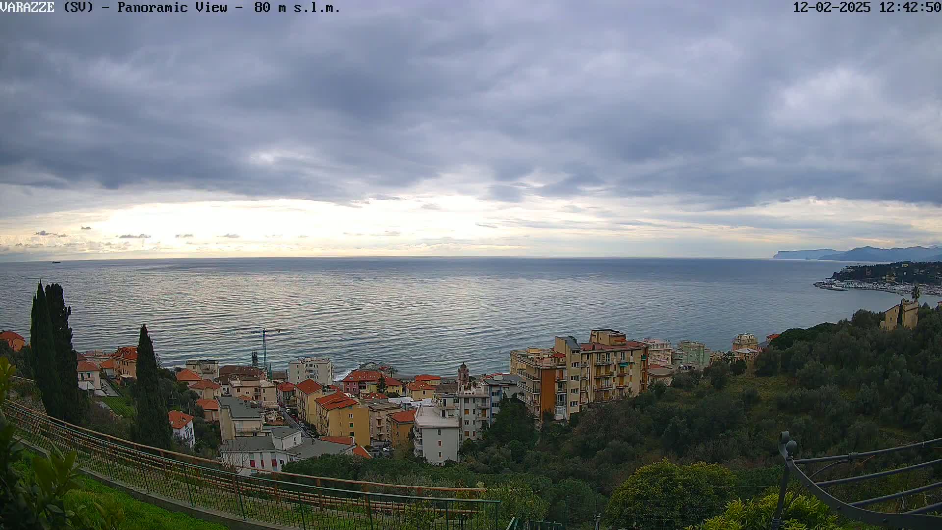 A coastal town with multi-story buildings overlooks a calm sea under a mostly cloudy sky.