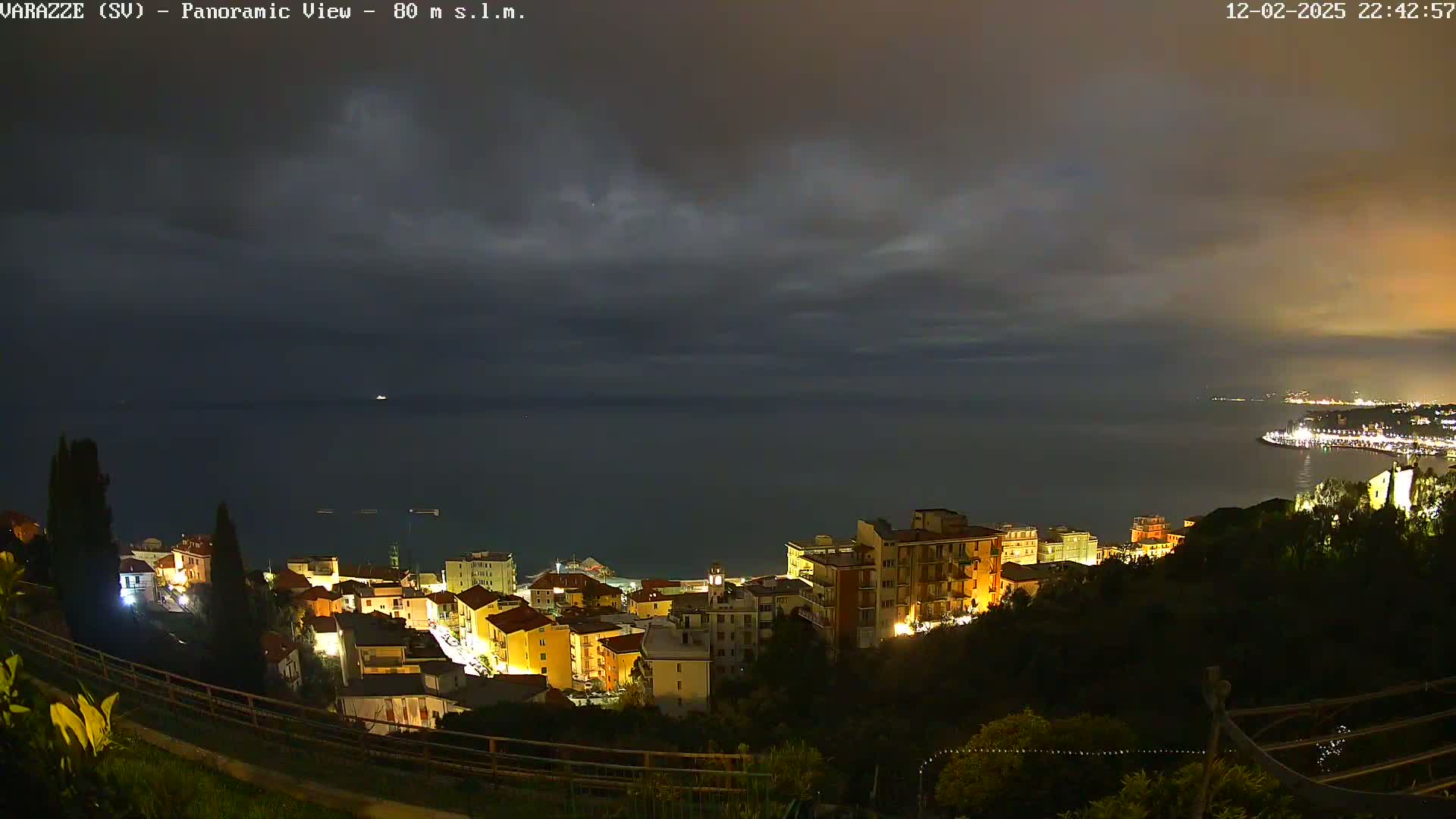 A nighttime panoramic view shows a coastal town illuminated against a dark, cloudy sky and a calm sea.