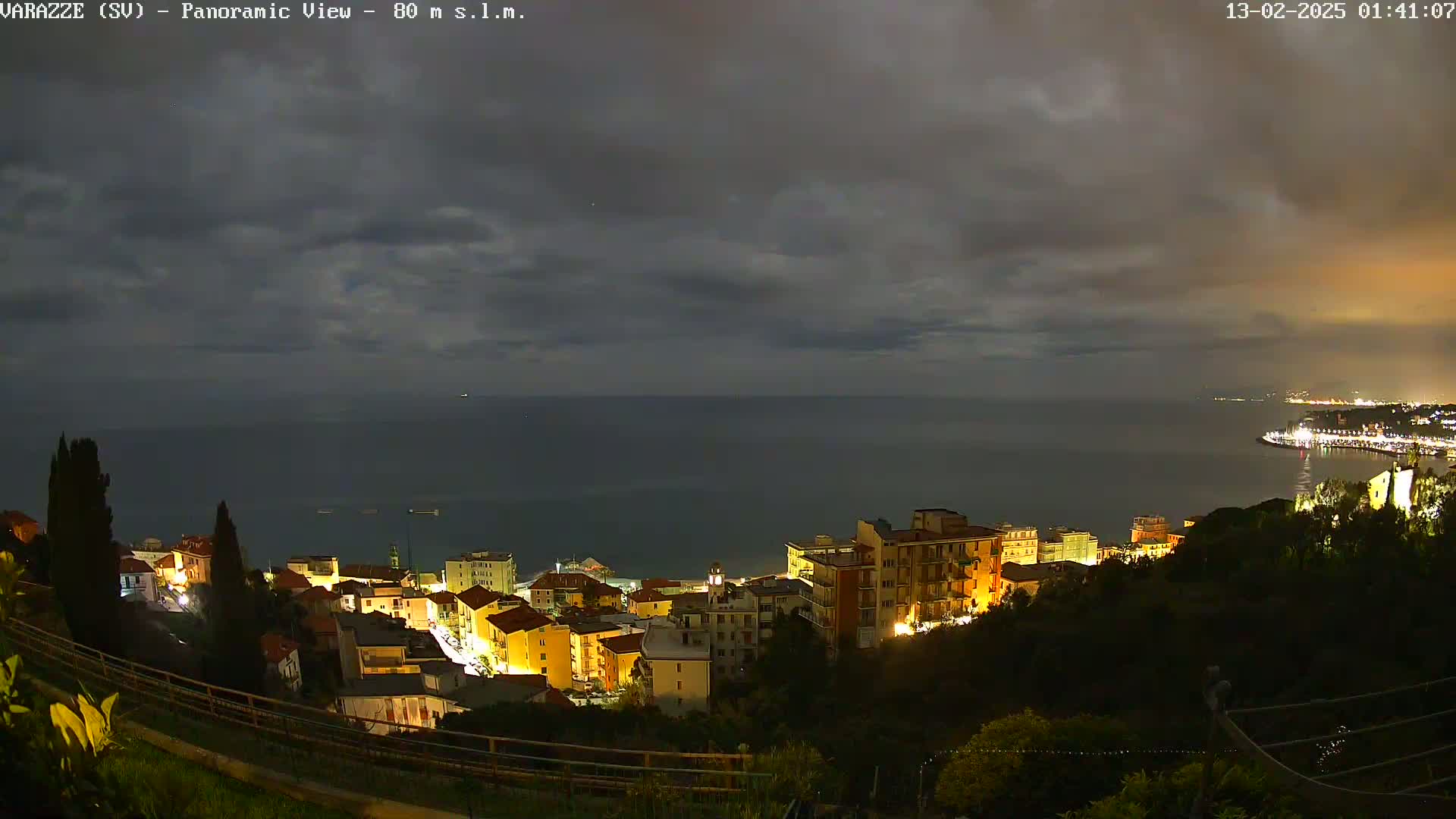 A nighttime, panoramic view of a coastal town illuminated with lights, under a cloudy sky, overlooking a dark sea.