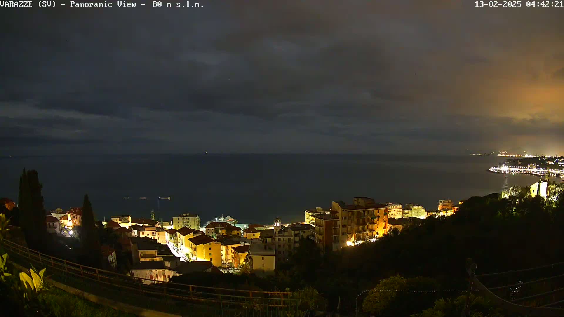 A nighttime panoramic view shows a coastal town with illuminated buildings overlooking a dark sea under a cloudy sky.