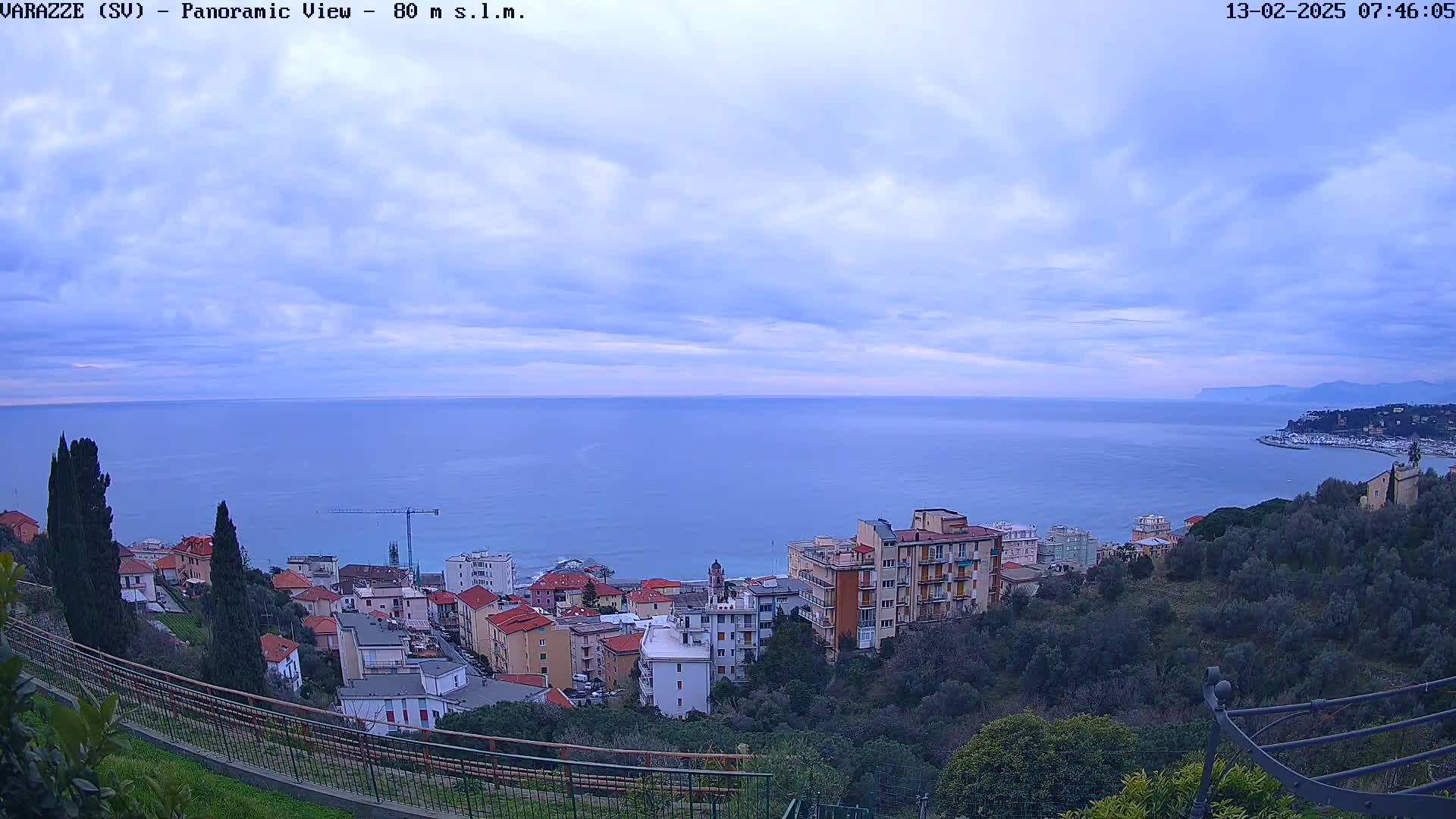 A coastal town with multi-story buildings overlooks a calm sea under a mostly cloudy sky.